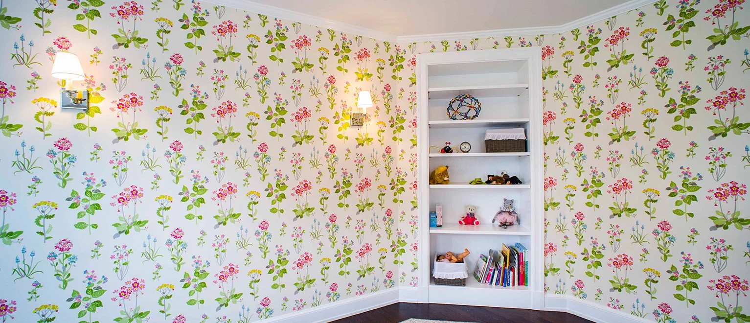 A corner of a room with floral wallpaper featuring pink, purple, and yellow flowers with green leaves, and two wall-mounted lamps with white shades. There is a white built-in bookshelf with stuffed animals, books, and decorative items.