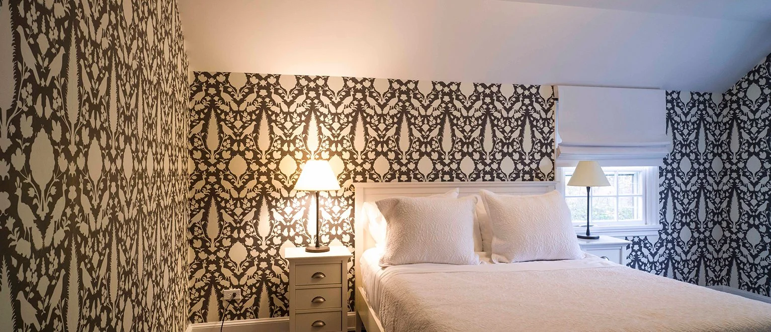A bedroom with patterned black and white wallpaper, a white bed with pillows, and two nightstands with lamps, one near the bed and one by a window with a white window shade.
