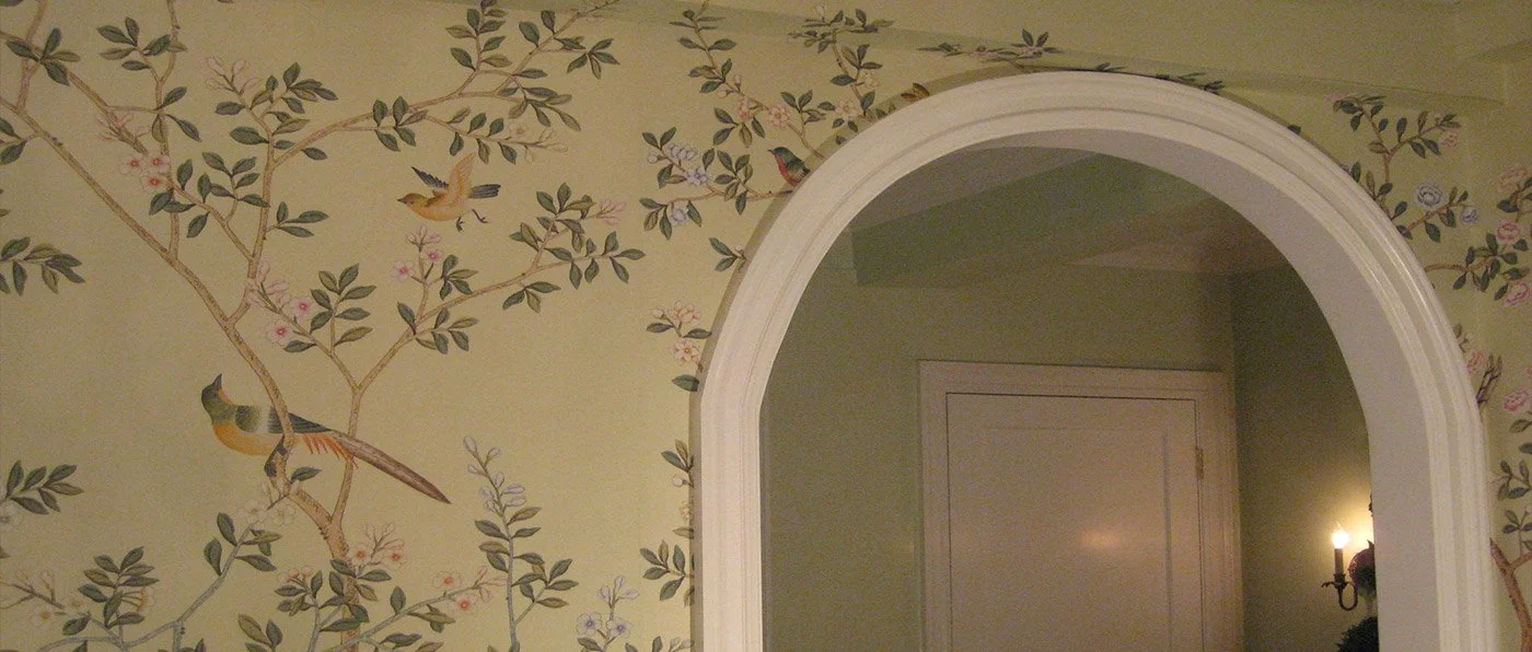 Wall with wallpaper featuring floral and bird design, and an arched doorway leading to a room with a closed door and wall sconce.