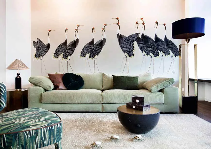 Living room with a light green sofa, decorative pillows, multiple lamps including a green patterned armchair, a round black coffee table, a beige rug, and a wall mural of stylized cranes.