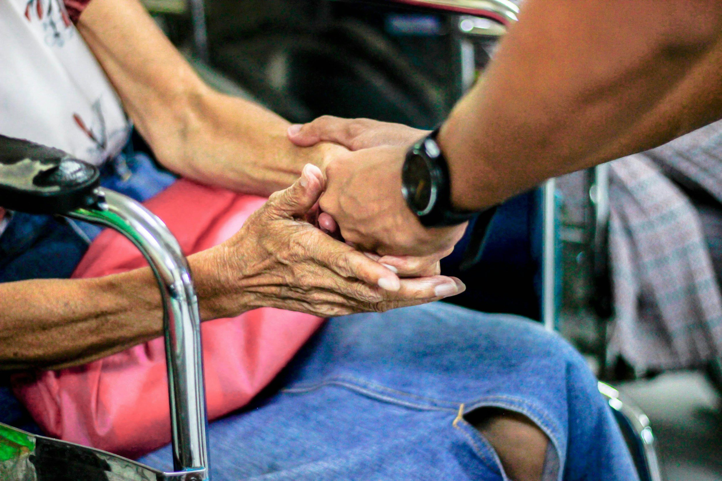 Two people, one elderly and one younger, holding hands in a comforting gesture. The elderly person is seated in a wheelchair, suggesting a caring or supportive moment in a public setting.