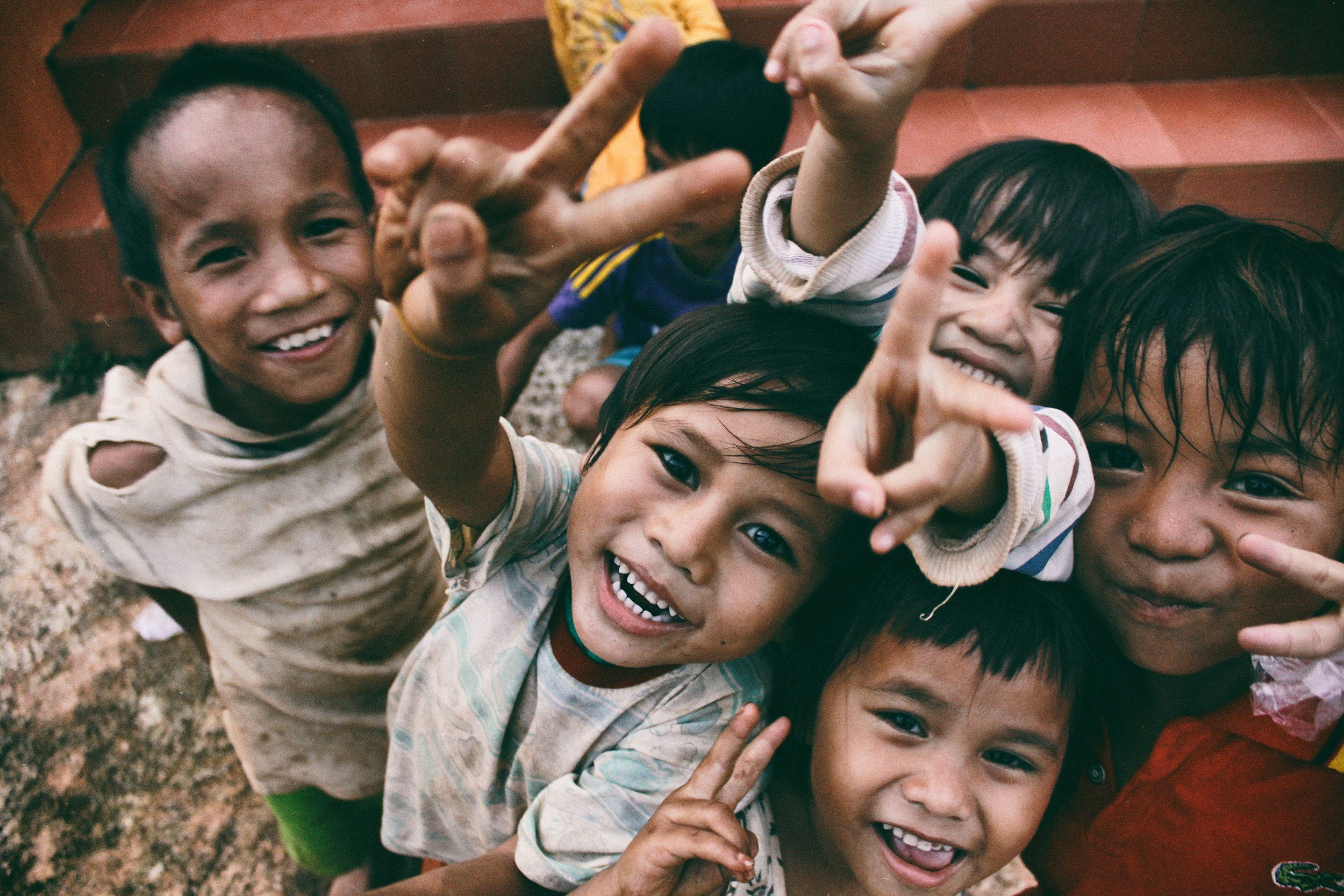 A group of children smiling, laughing, and playing outdoors, with some making peace signs and others reaching towards the camera.