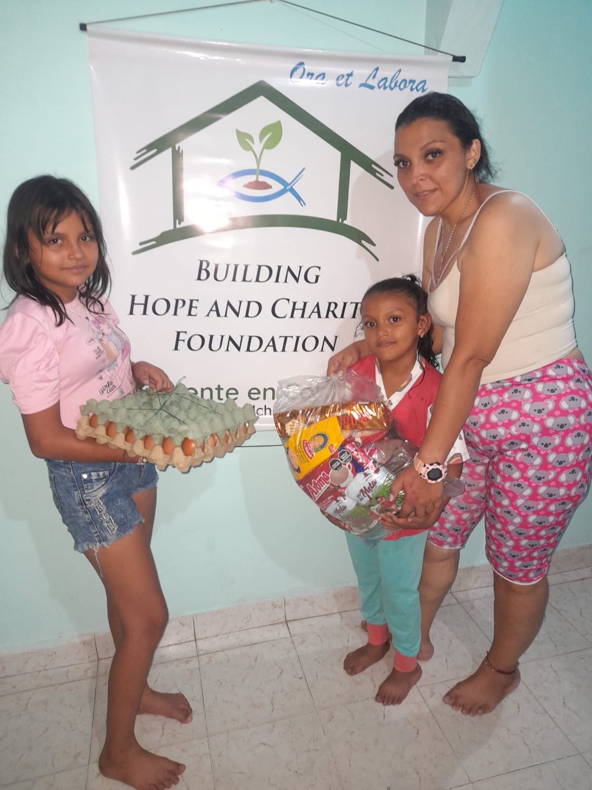 A woman and two young girls standing indoors in front of a white banner that reads 'Building Hope and Charity Foundation'. The woman is handing a bag of supplies to one girl and the other girl is holding a tray of eggs.
