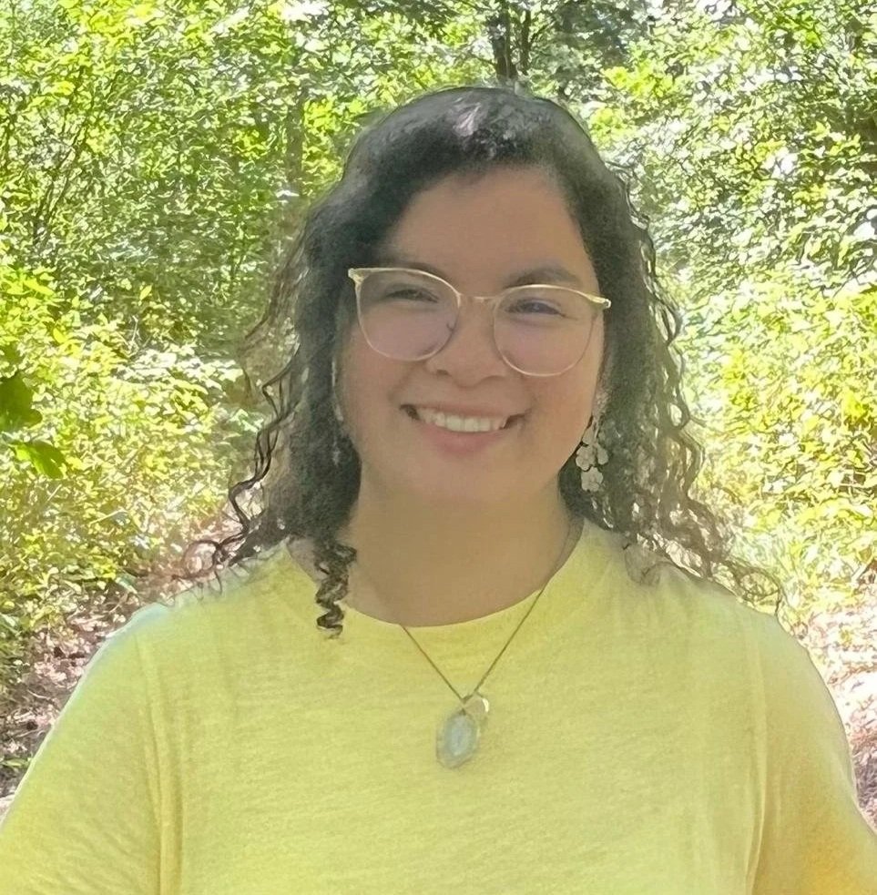 A woman with curly dark hair, light skin, and glasses, smiling outdoors with green foliage in the background, wearing a yellow top and a necklace.