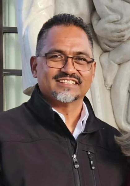 A smiling man with glasses, short dark hair with gray, and a goatee, wearing a black jacket, standing in front of a white stone sculpture, with a window and curtain in the background.