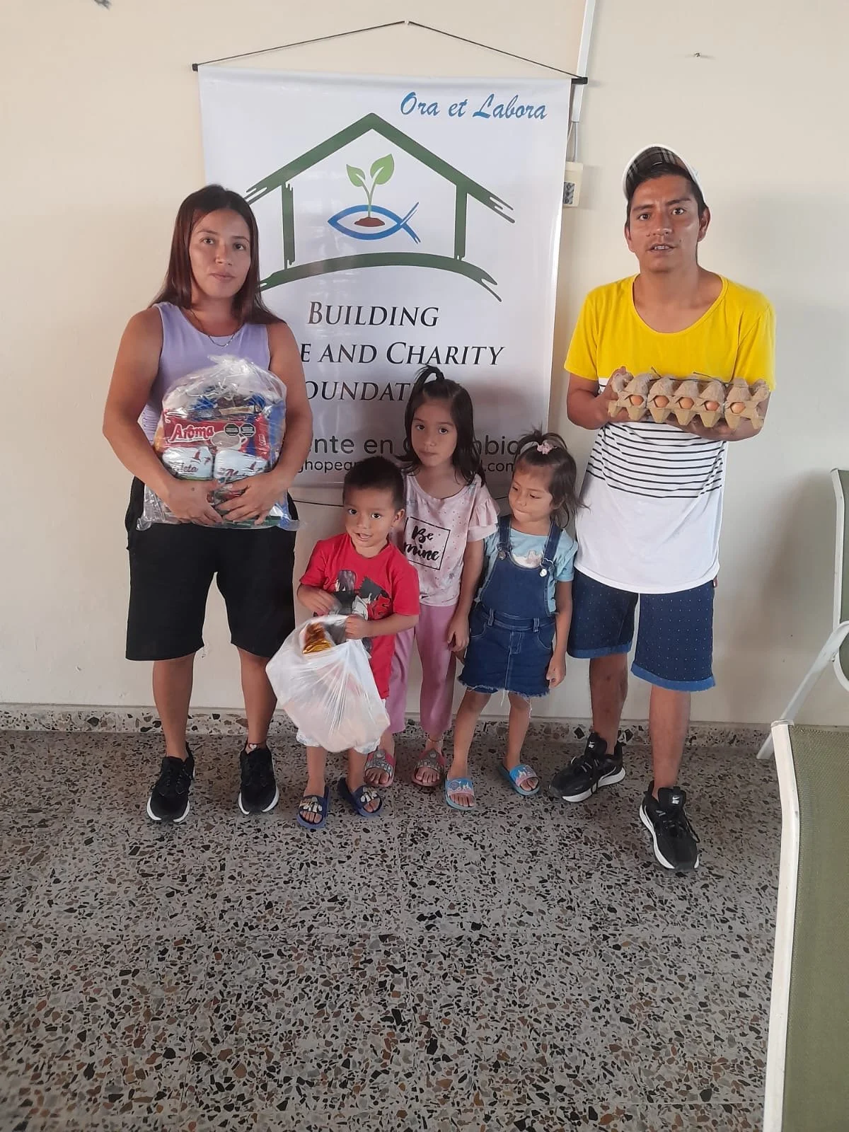 People standing in front of a charity banner, holding food and supplies, with children and adults smiling.