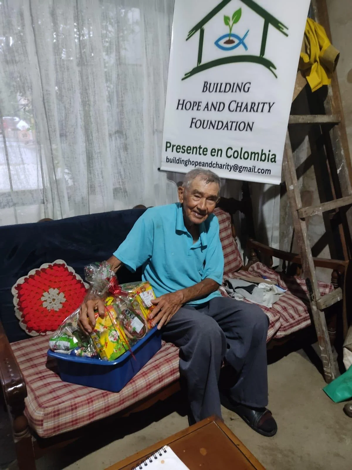 An elderly man sitting on a wooden bench in front of a white banner with green, blue, and black text. He is holding a gift basket wrapped in plastic with various snacks and treats. The man is smiling, wearing a light blue shirt and dark pants, with a crochet pillow and blanket behind him. The setting appears to be indoors with a transparent curtain in the background and a wooden ladder on the right side.