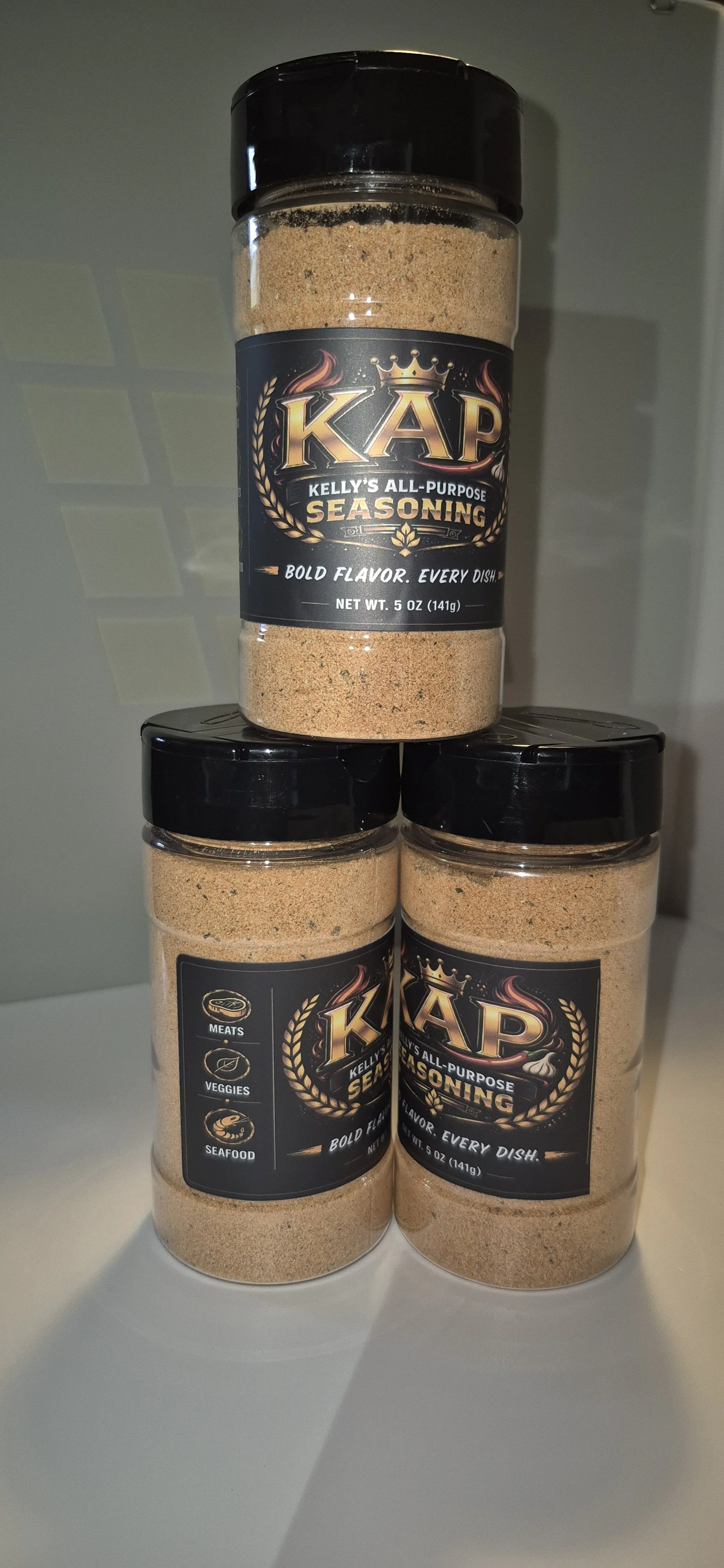 Three containers of Kelly's All-Purpose Seasoning stacked on a white surface, with the top container showing the seasoning and the two below displaying labels indicating it can be used for meats, veggies, and seafood.