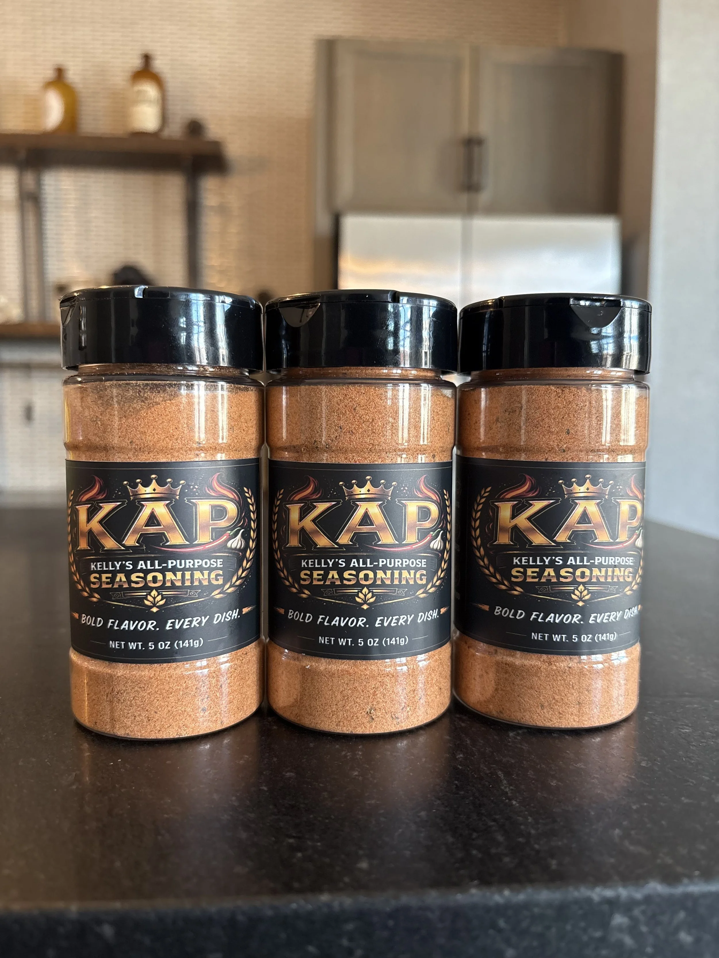 Three bottles of Kelly's All-Purpose Seasoning on a dark countertop, with a blurry background of kitchen cabinets and wall decor.