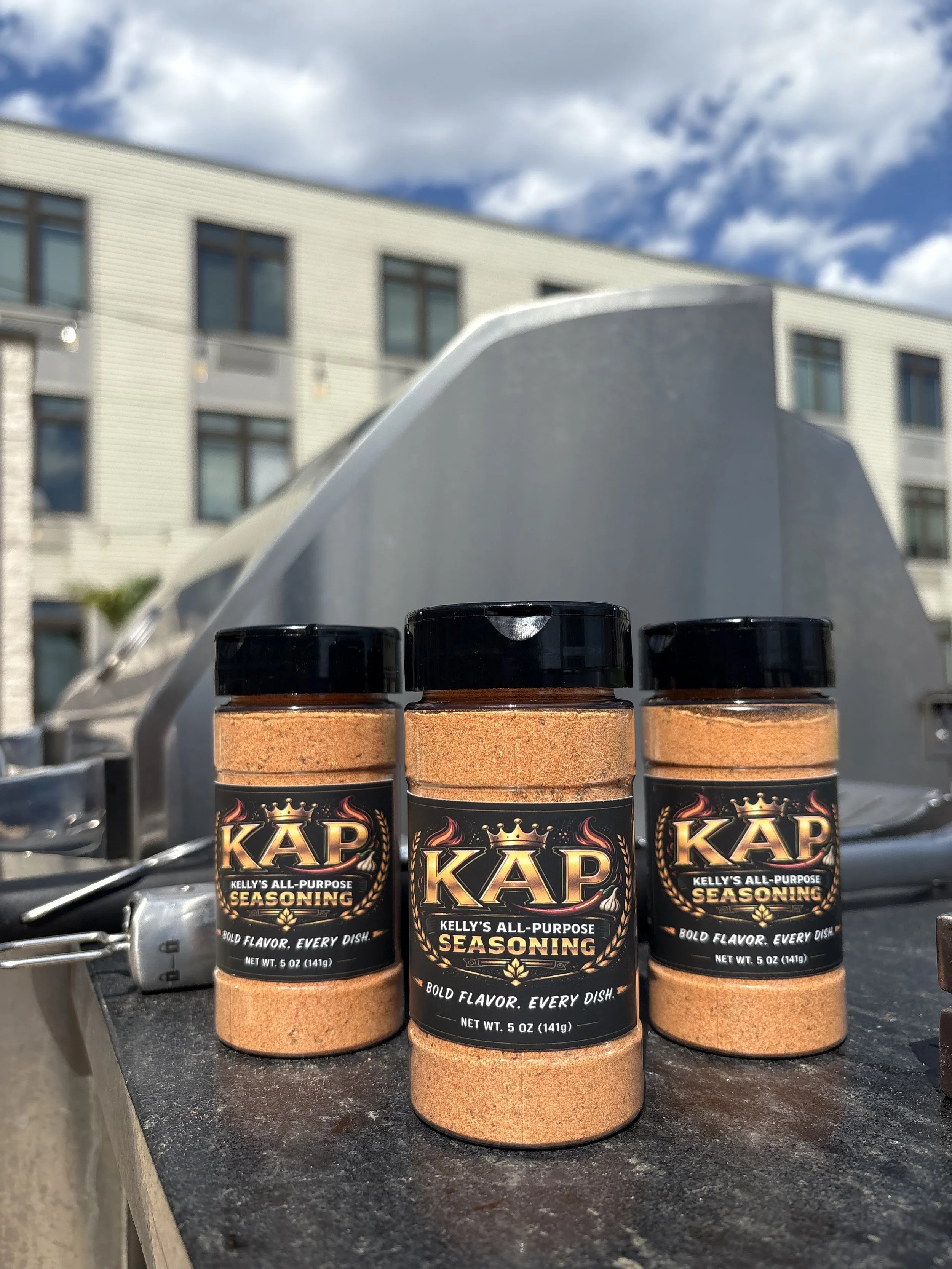 Three bottles of Kelly's All-Purpose Seasoning placed on a black surface in front of a building with multiple windows, under a partly cloudy sky.