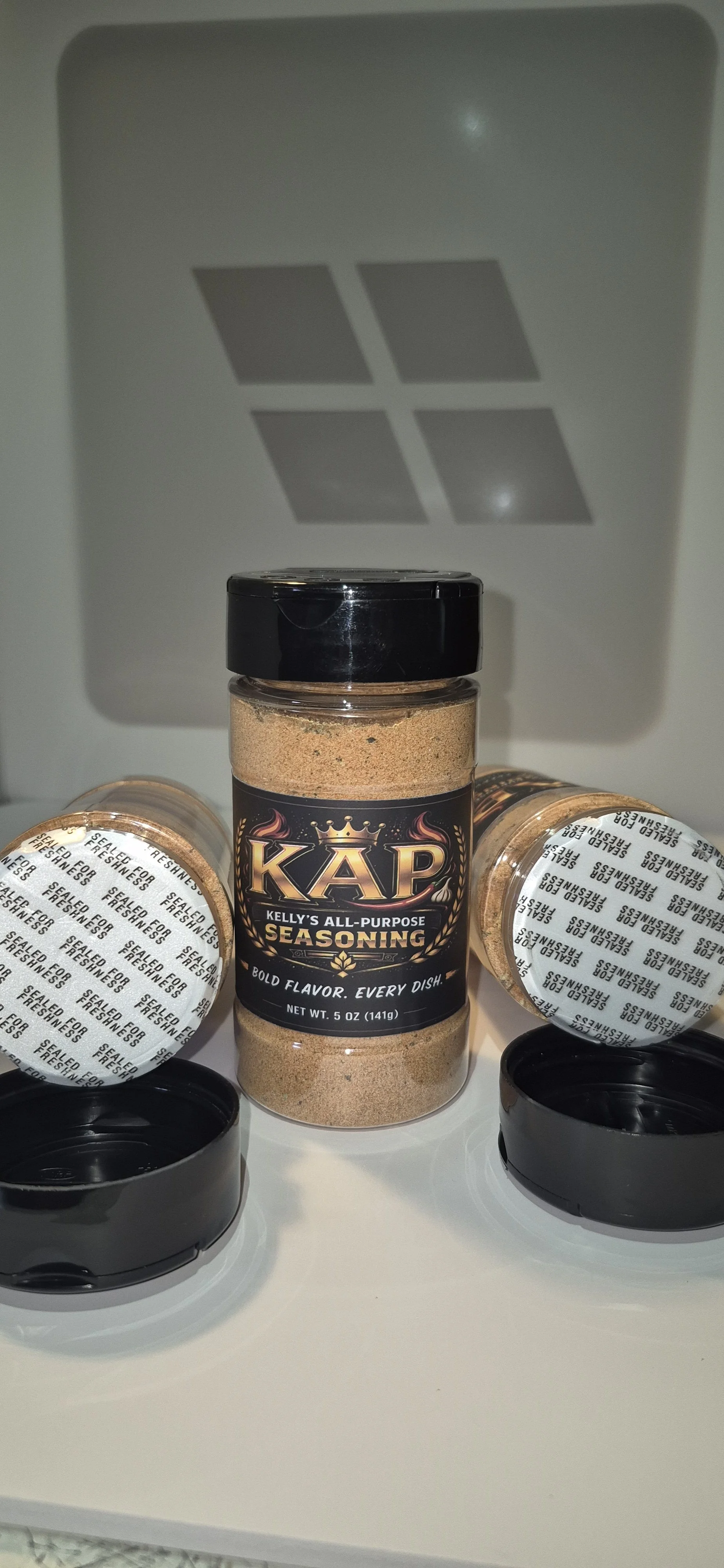 A jar of Kelly's All-Purpose Seasoning in front of a pantry or appliance with a logo, surrounded by other seasoning jars. The seasoning jar has a black lid and black label with gold and white text.
