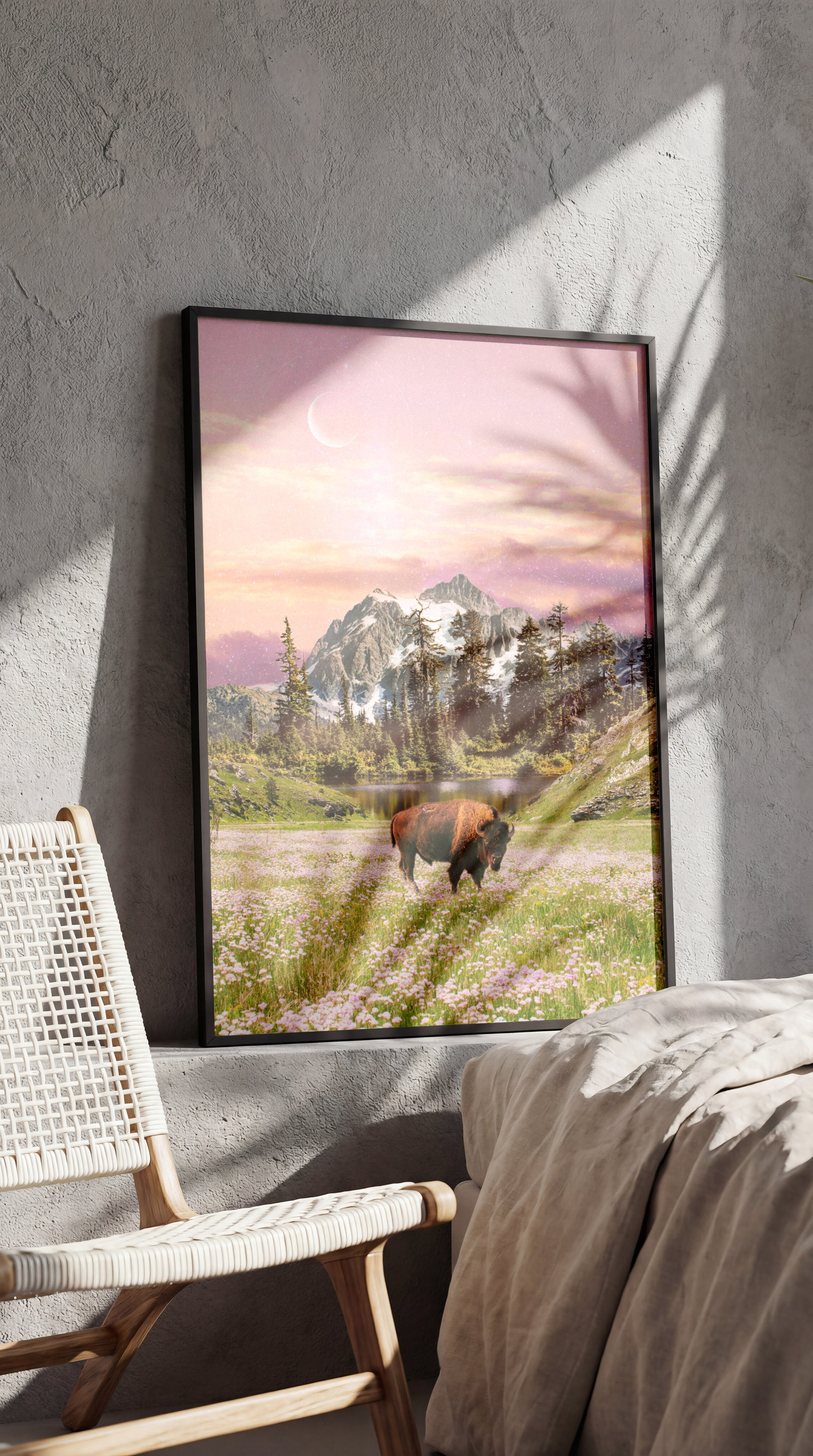 Framed landscape painting of mountains, trees, a river, and a bison in a meadow, with a pink and purple sky and a crescent moon, leaning against a textured wall near a bed and a wicker chair.