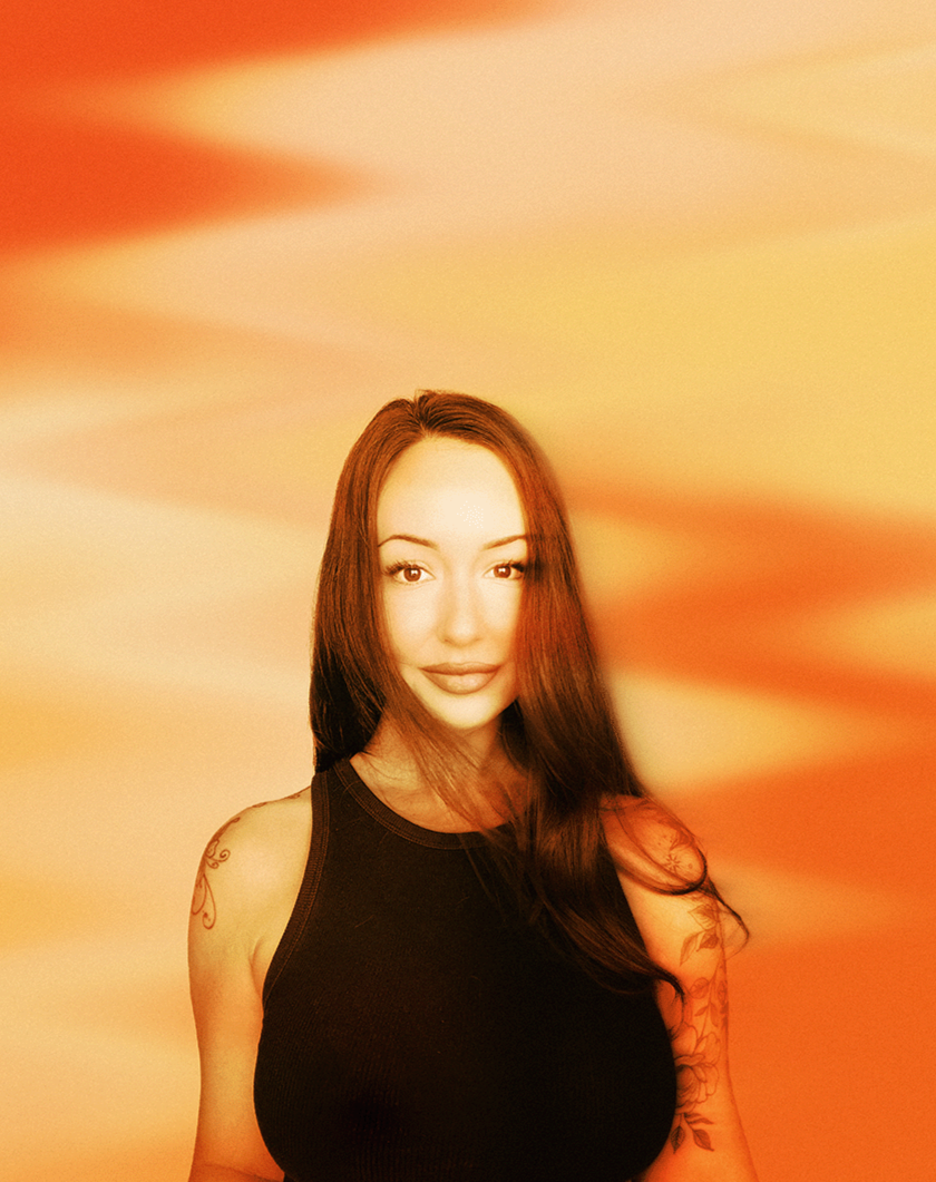 A woman with long, reddish-brown hair and tattoos on her arms, wearing a black sleeveless top, stands against a colorful, blurred sunset background.