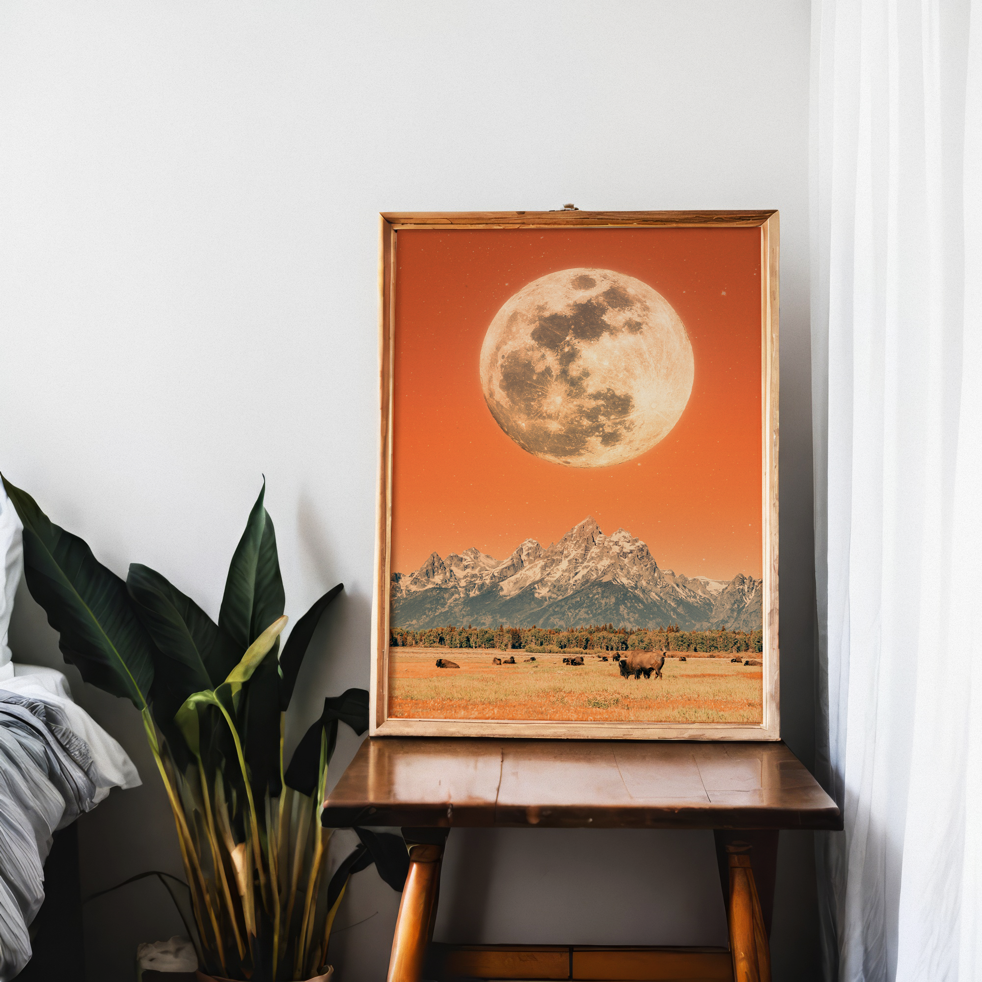 A large artwork of the moon on a red-orange background lies inside a wooden frame on a desk. In front of it, there is a mountain landscape with buffalo grazing in a field. To the left, there's a green leafy plant and a neatly made bed.