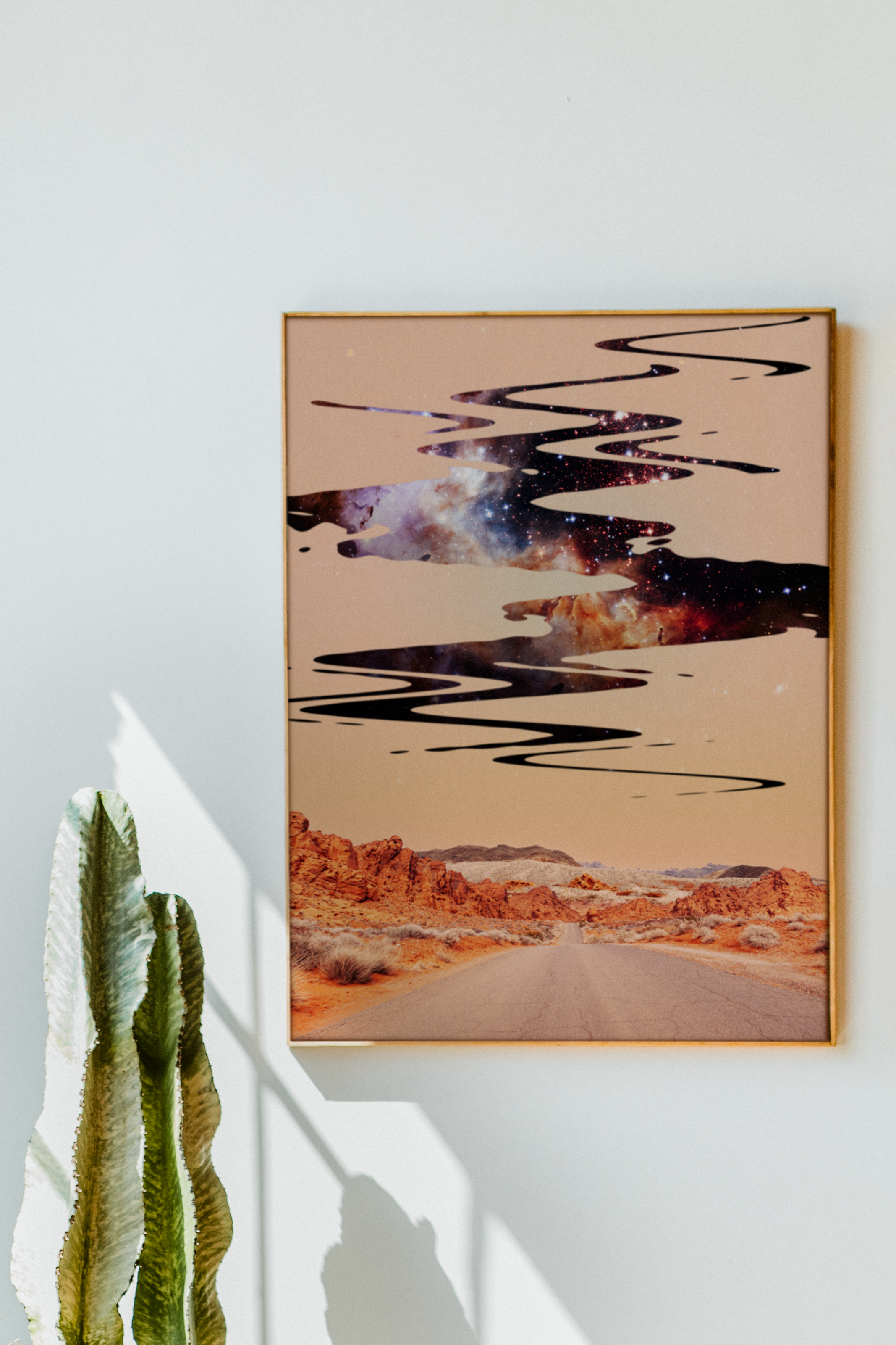 Decorative wall art showing a desert landscape with rocky formations and a galaxy sky, with black paint streaks across, and a tall cactus in the foreground casting a shadow on a white wall.