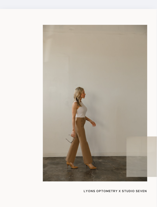 A woman walking in profile against a plain white wall, wearing a white sleeveless top, beige wide-leg pants, and yellow high heels, holding glasses in her right hand.