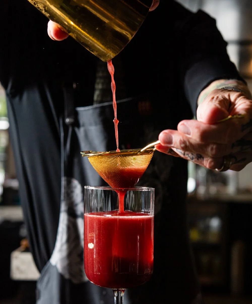 Person pouring red liquid through a strainer into a glass.