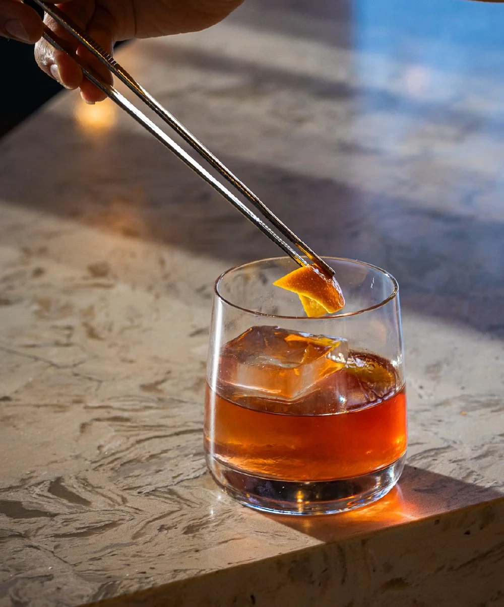 A hand holding metal tongs with an orange peel over a glass of amber-colored cocktail with ice cubes on a textured surface.