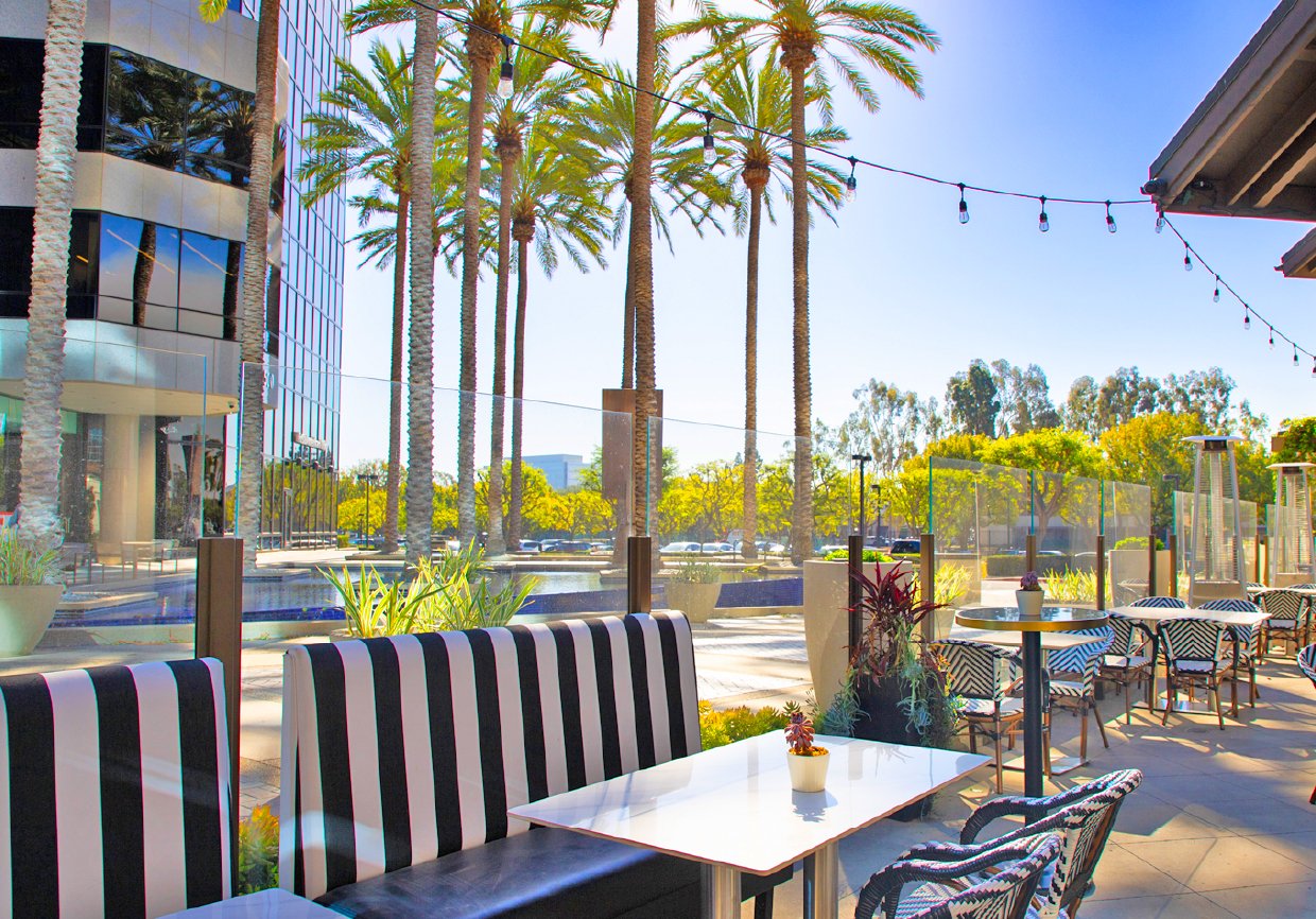 Outdoor patio seating area at a restaurant with striped chairs, small tables with plants, and glass partitions. Tall palm trees and a modern glass building are visible in the background on a sunny day.