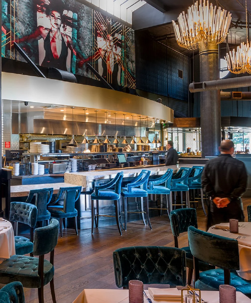 Interior of a modern restaurant with a bar area, blue velvet chairs, and a kitchen in the background. Decorative chandeliers hang from the ceiling, and a large artwork features a stylized female portrait.