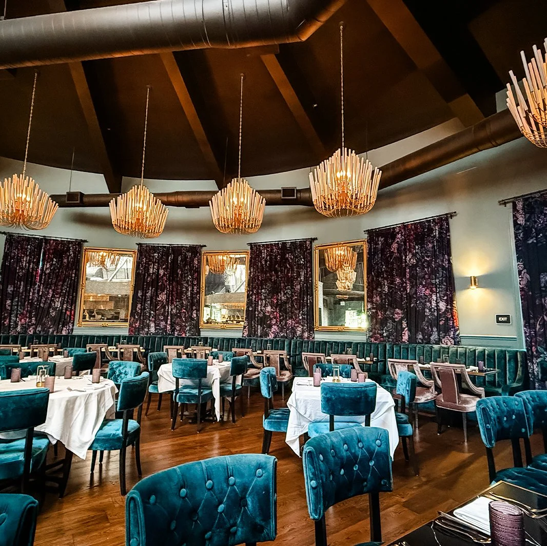Elegant restaurant interior with round tables covered in white tablecloths, teal and purple upholstered chairs, dark floral drapes, mirrors on the wall, and large wooden chandeliers hanging from a dark wooden ceiling.