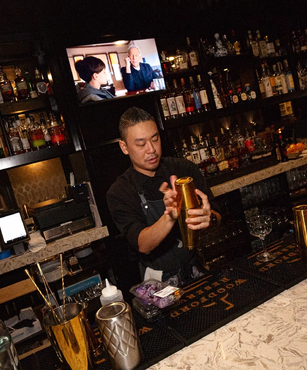 A bartender preparing a cocktail behind a bar with various liquor bottles. A TV screen displays a scene with two people, one on the phone. The bar has a marble countertop and bar tools.