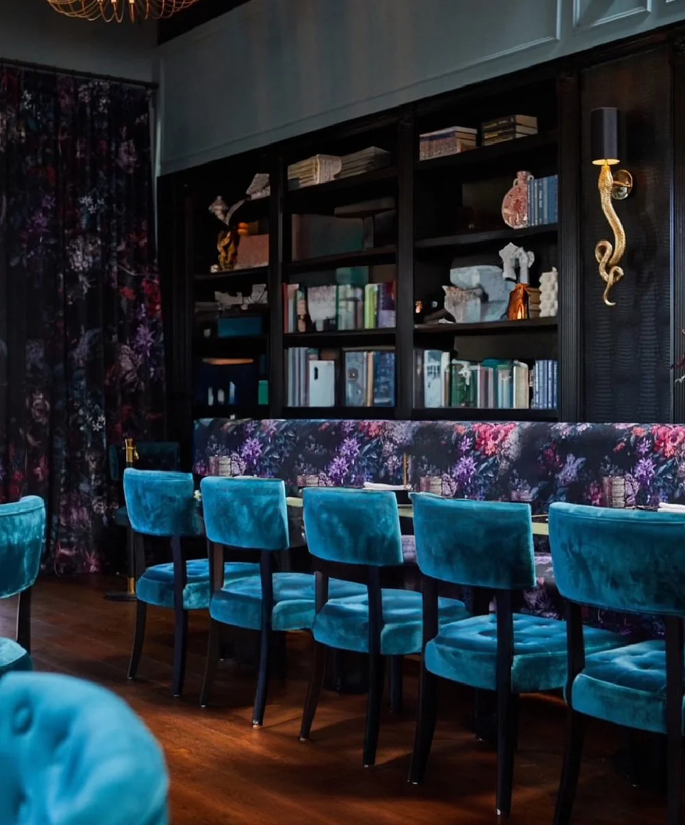 Interior of a stylish restaurant with a long floral-patterned upholstered banquette, teal velvet chairs, dark wood floors, and a black bookshelf with decorative objects and books on a dark wall.