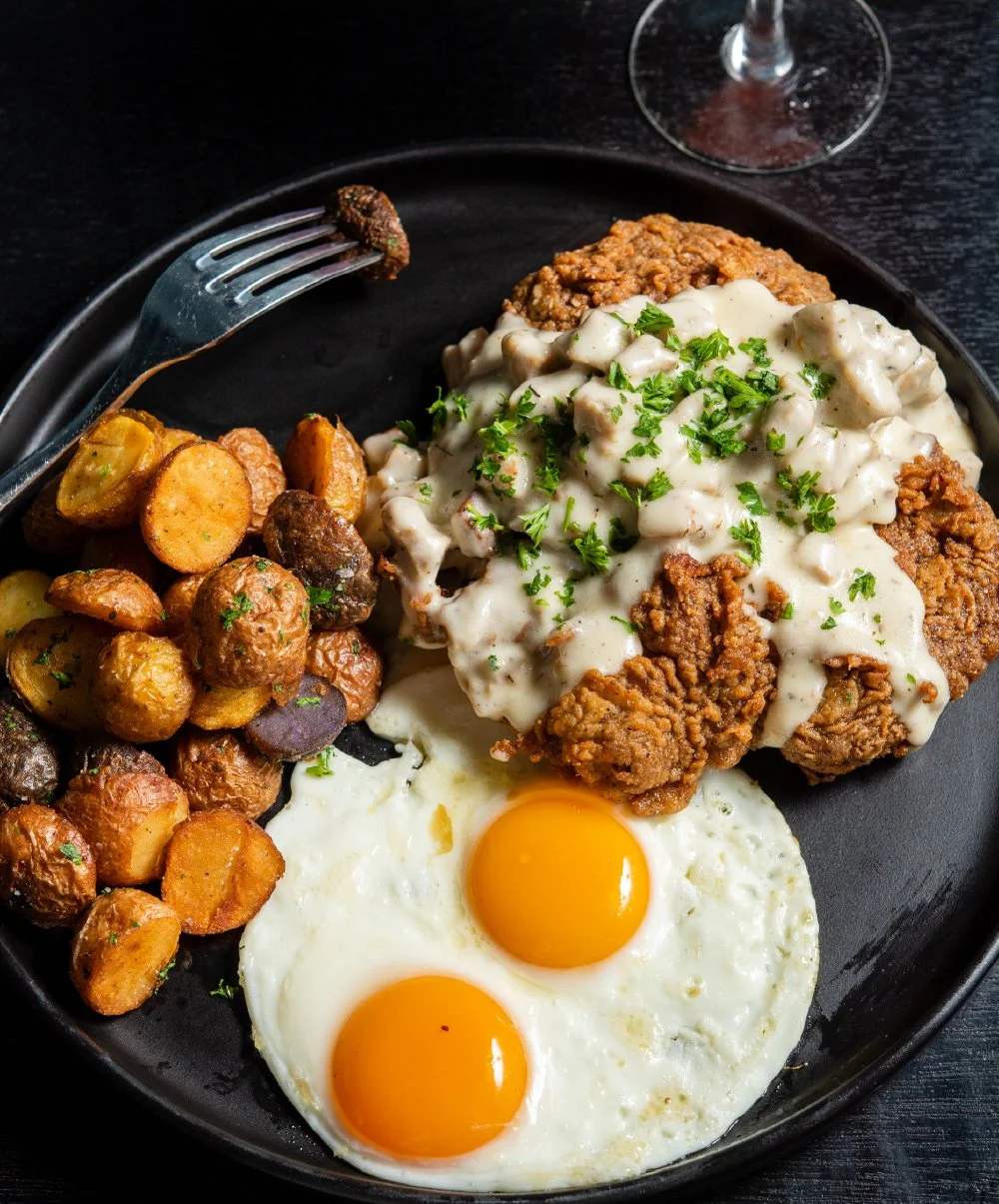 Fried chicken with white gravy, two sunny side up eggs, roasted potatoes, and a glass of wine on a black plate.