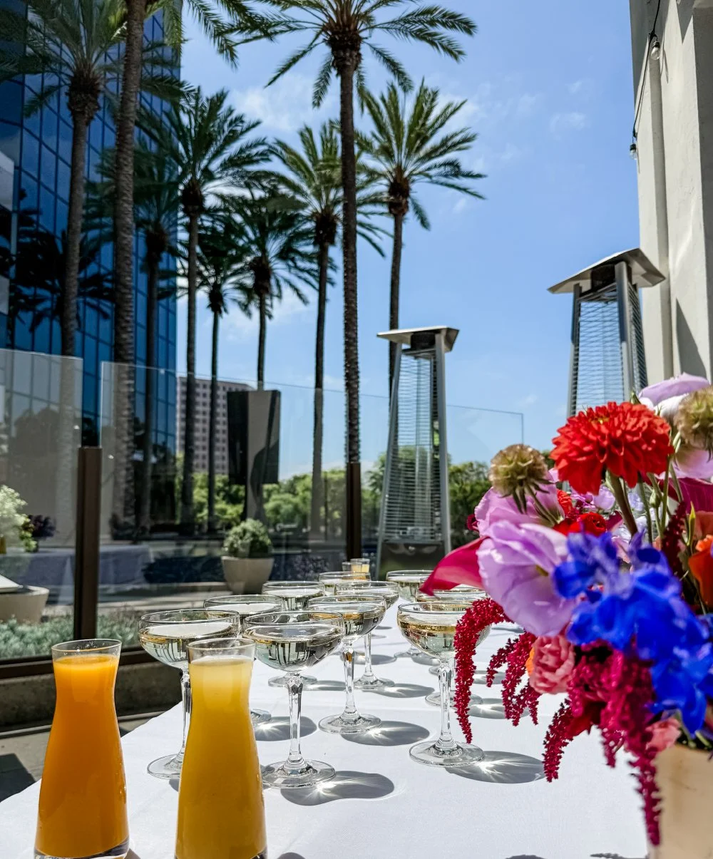 Outdoor event setting with tall palm trees, colorful flower arrangement, glasses of champagne, and two carafes of orange juice on a white tablecloth, with a cityscape in the background.