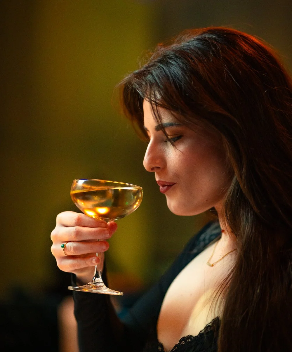 A woman with dark hair holding a glass of white wine and smelling it, with a blurred dark background.