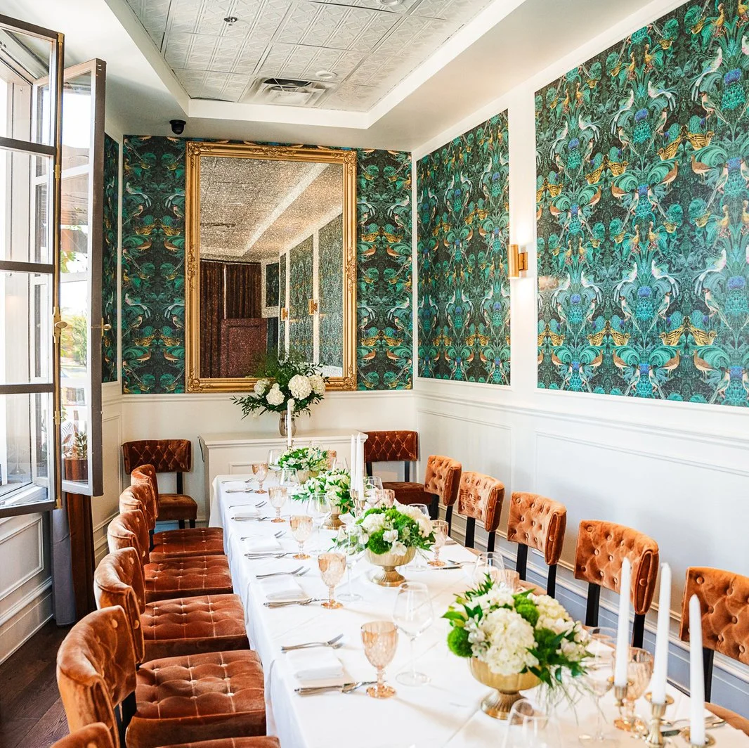 A dining room set for a formal event with a long white table, peach velvet chairs, floral centerpieces, candles, and a large mirror on the wall. The room has tropical patterned wallpaper and a window letting in natural light.