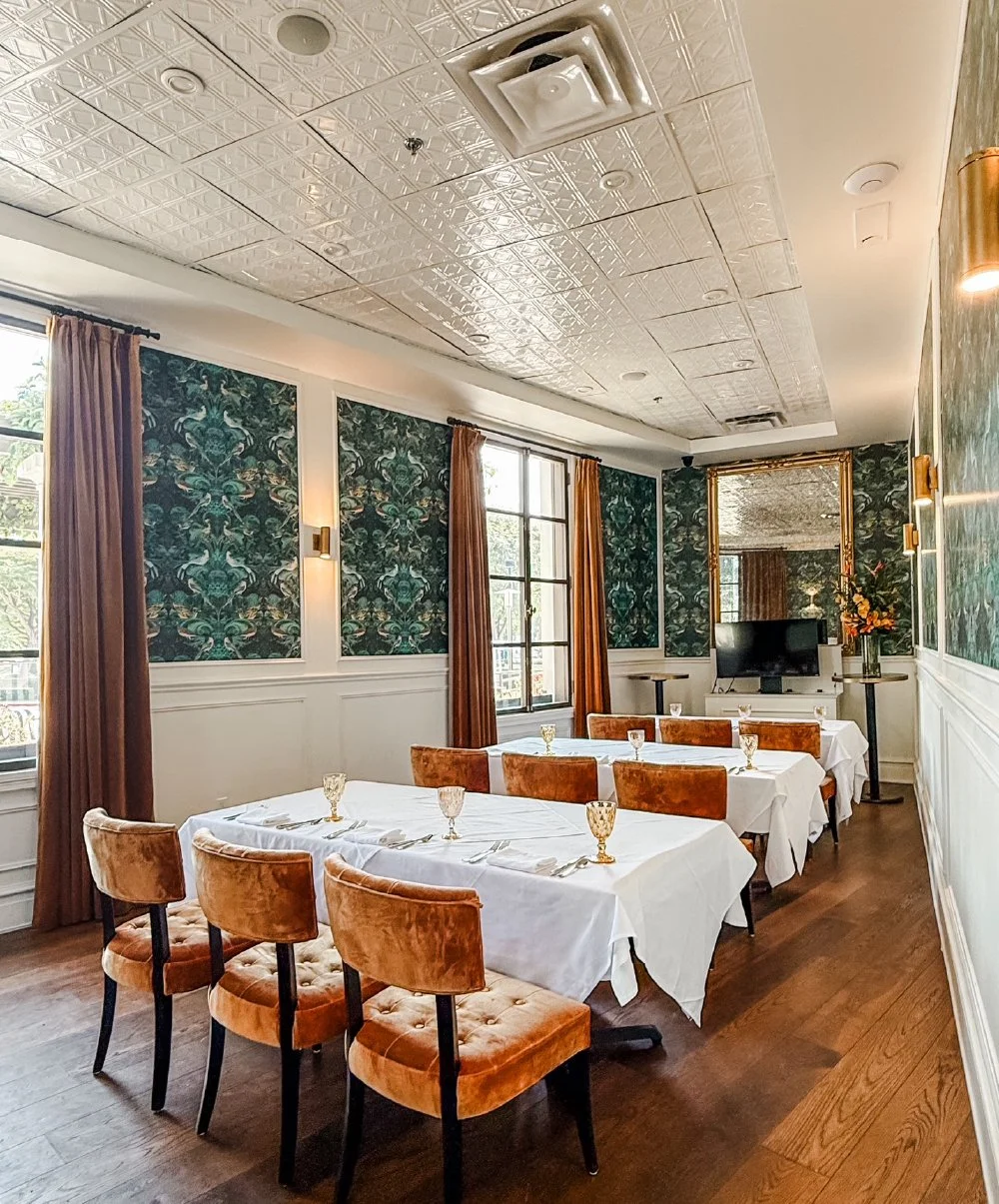 A dining room with two rectangular tables covered in white tablecloths, set with gold-rimmed glasses and silverware. Eight orange velvet chairs are placed around the tables. The room has patterned dark wallpaper, large windows with brown curtains, an