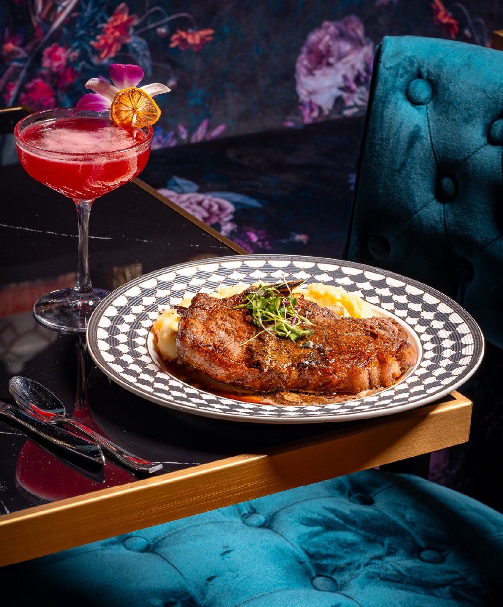 A plate of grilled steak topped with greens, served with mashed potatoes on a decorative plate, accompanied by a pink cocktail garnished with flowers and a dried citrus slice, set on a black table with a teal cushioned chair and floral wallpaper back