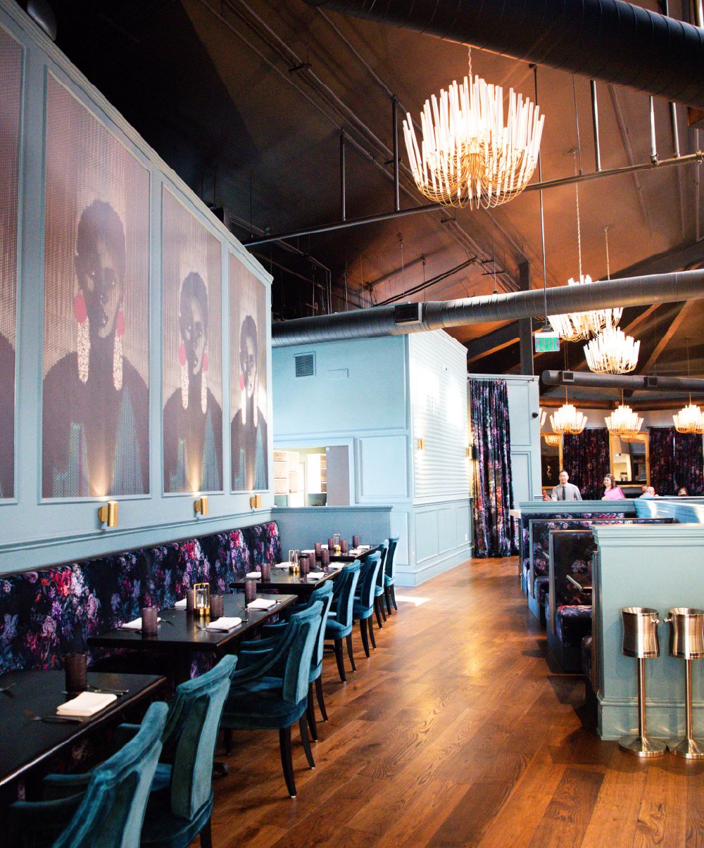 Interior of a stylish restaurant with a row of tables and chairs, floral upholstery, and modern chandeliers on a wooden floor. Large portraits of a woman with earrings and earrings are displayed on the wall, with a partially visible standing person i
