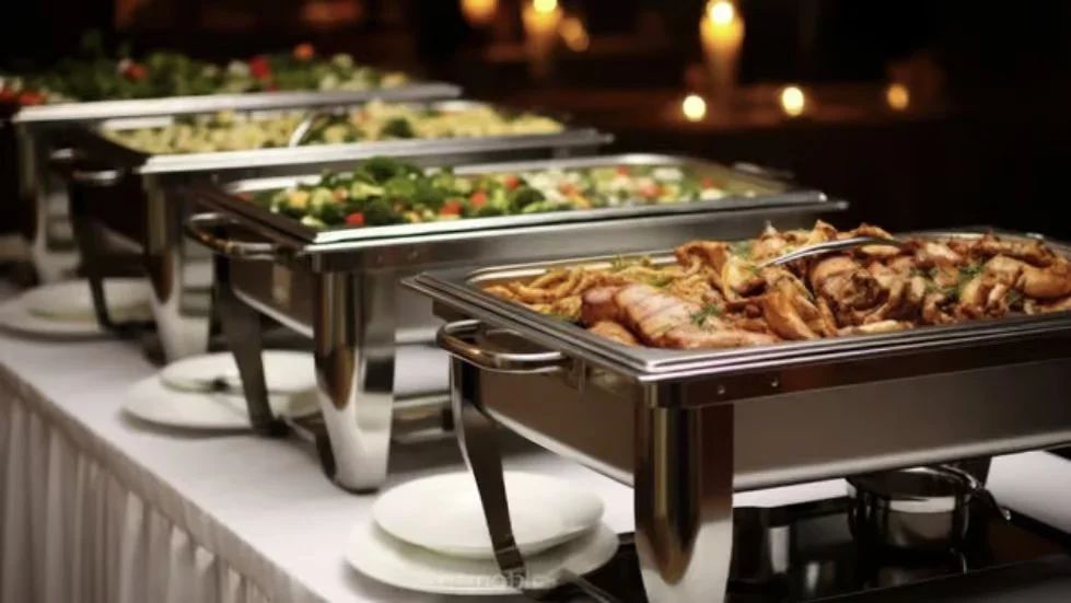 Buffet table with chafing dishes containing salad, cooked chicken, and other dishes, set with plates and serving utensils.