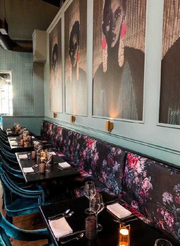 A restaurant interior with a long table set for dining, featuring wine glasses, candles, and napkins. The wall has large artistic portraits of women with dark hair and pink earrings, and there are velvet chairs with floral patterns and others in teal