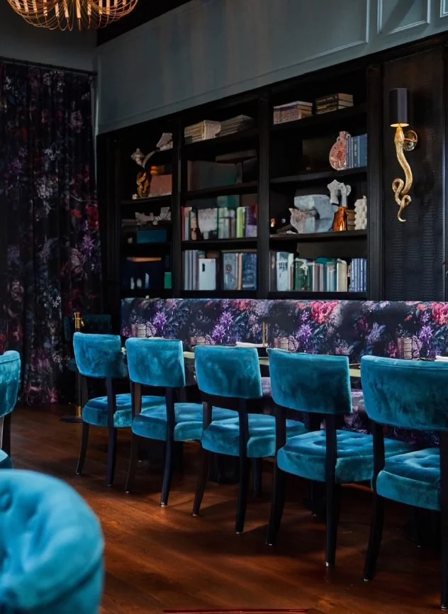 Interior of a stylish room with a dark wooden bookshelf filled with books and decorative objects, dark floral curtain, and a row of plush teal velvet chairs along a floral upholstered bench, with wooden flooring.