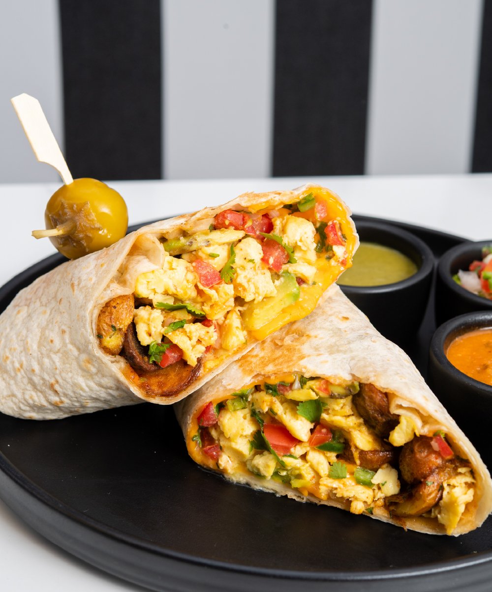 Two breakfast burritos filled with scrambled eggs, tomatoes, green peppers, and other ingredients on a black plate with small bowls of green and orange salsa in the background. A jalapeño pepper with a toothpick is placed on top of one burrito.