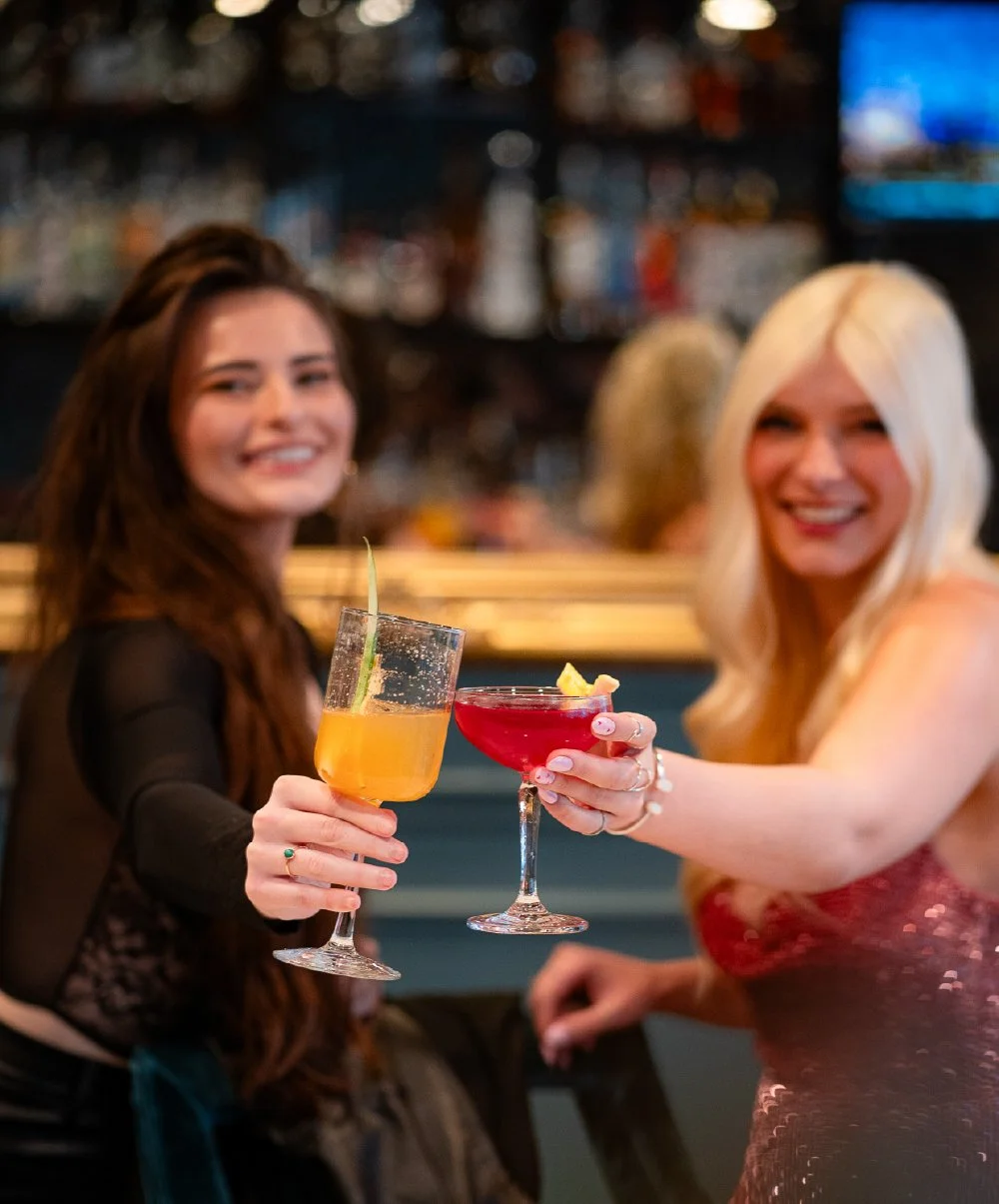 Two women clinking cocktail glasses at a bar, one with a yellow drink and the other with a red drink, both smiling and looking at the camera.
