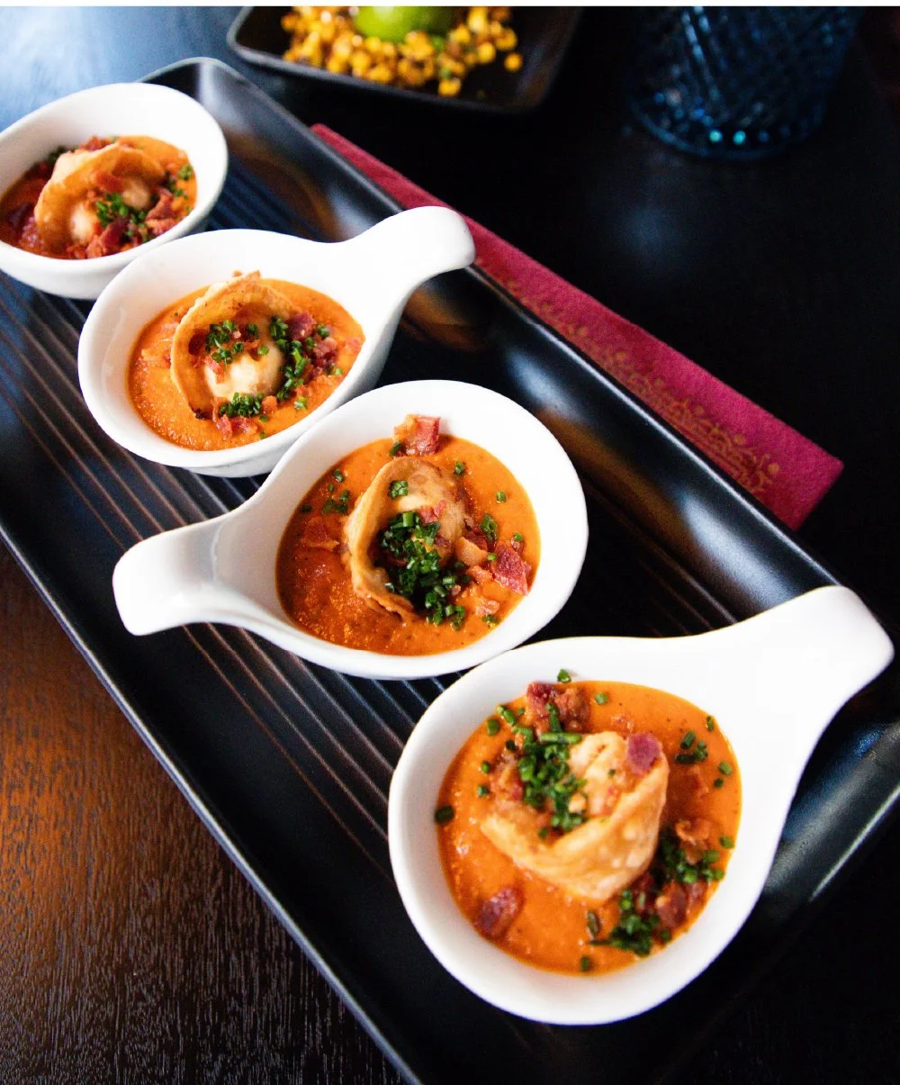 Four small white bowls with spoon-shaped handles containing stuffed bell peppers in tomato sauce, garnished with chopped green herbs, on a black tray.