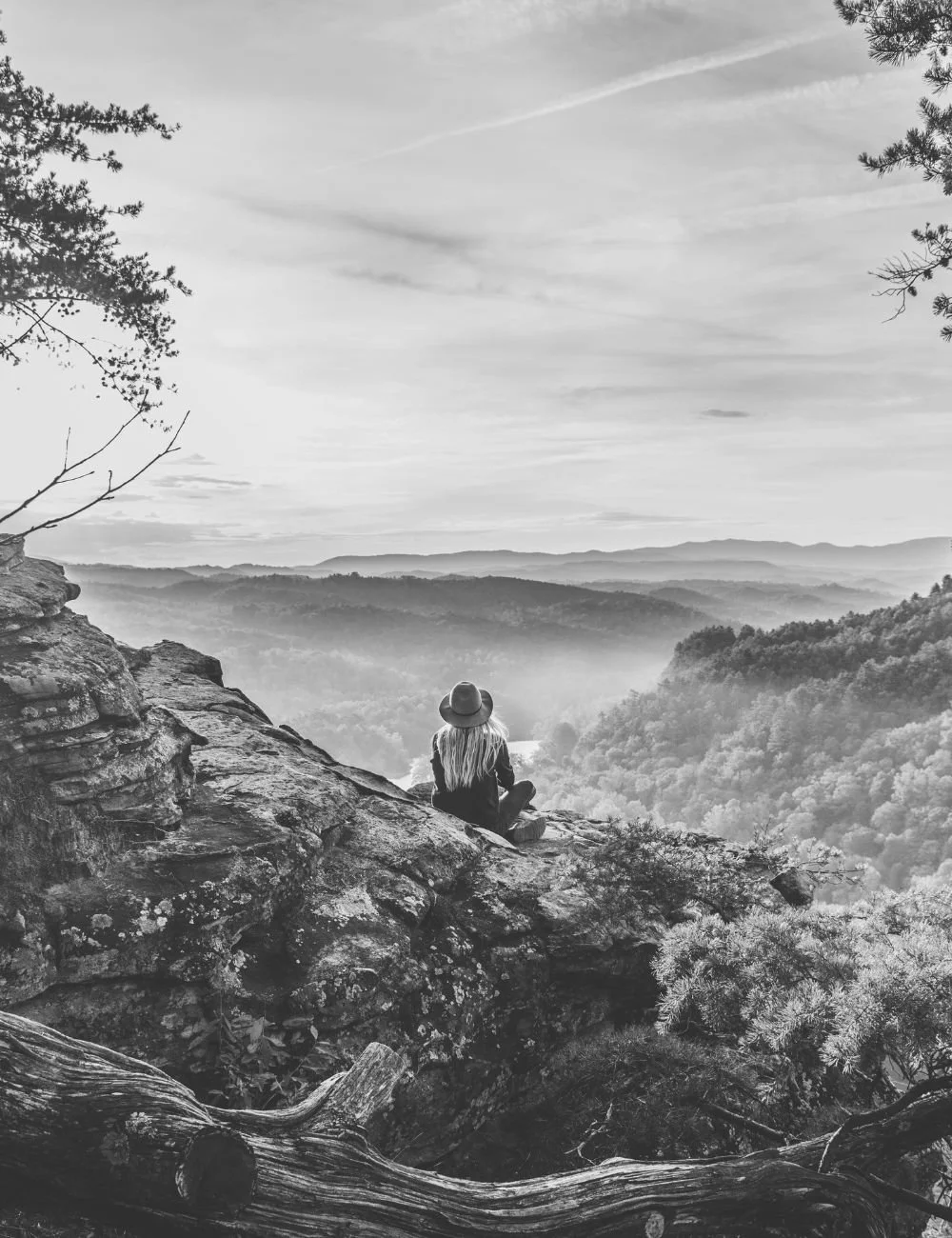 A woman with long hair, wearing a hat and sitting on a rocky ledge, overlooking a vast valley with rolling hills and trees, in a black and white photo.