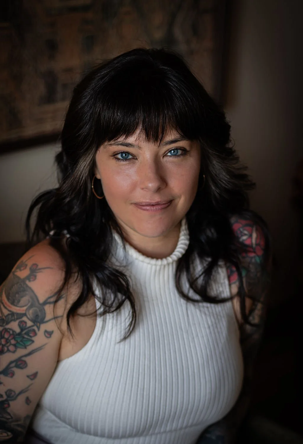 A woman with dark wavy hair, blue eyes, and tattoos on her arms, wearing a white sleeveless turtleneck top, smiling at the camera.