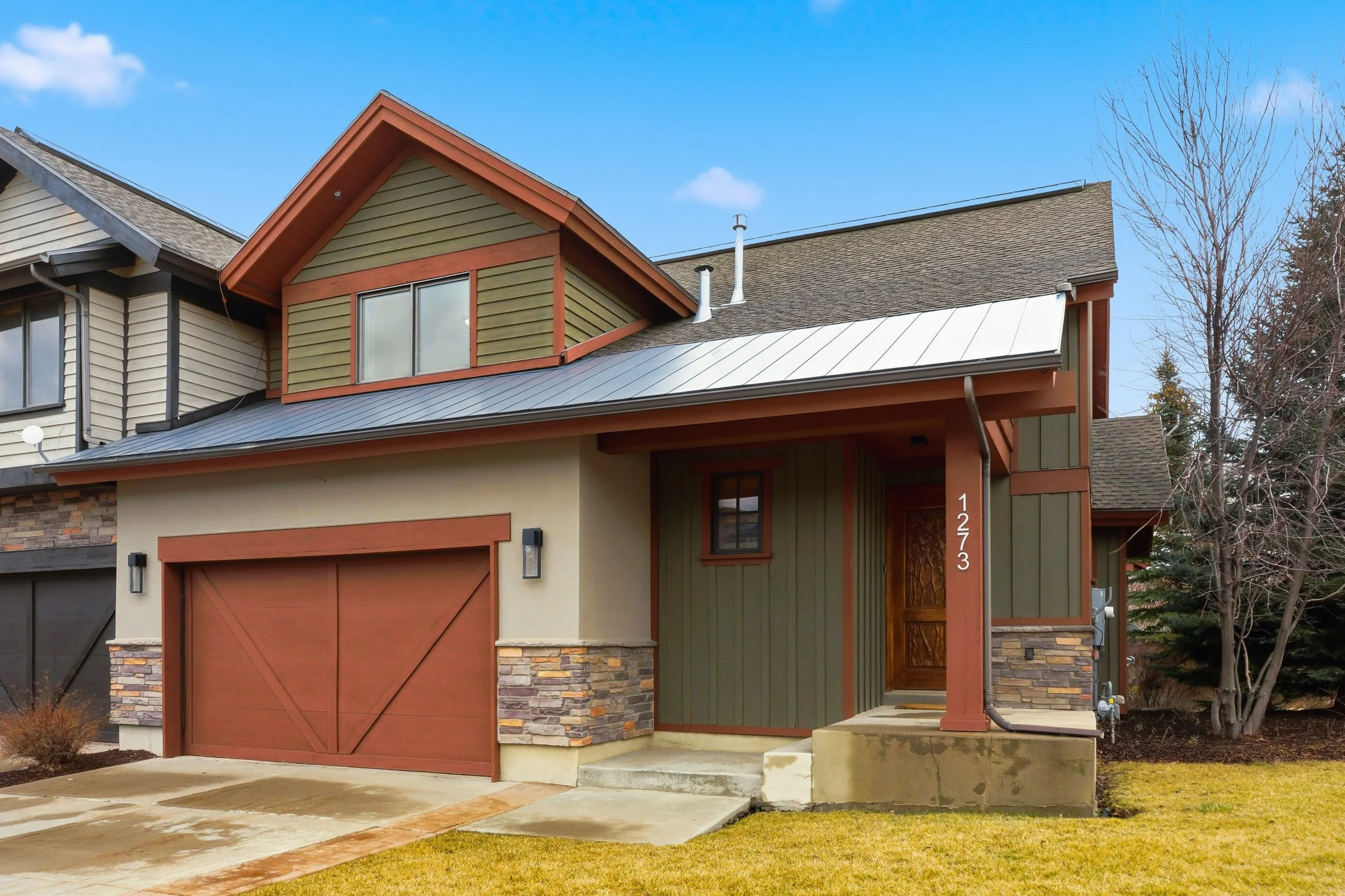 Front view of a modern house with a brown garage door, green and brown siding, stone accents, and a small porch. The house has a gabled roof with two chimneys and a window with a grid pattern. There is a small staircase leading to the front door and a well-maintained lawn.