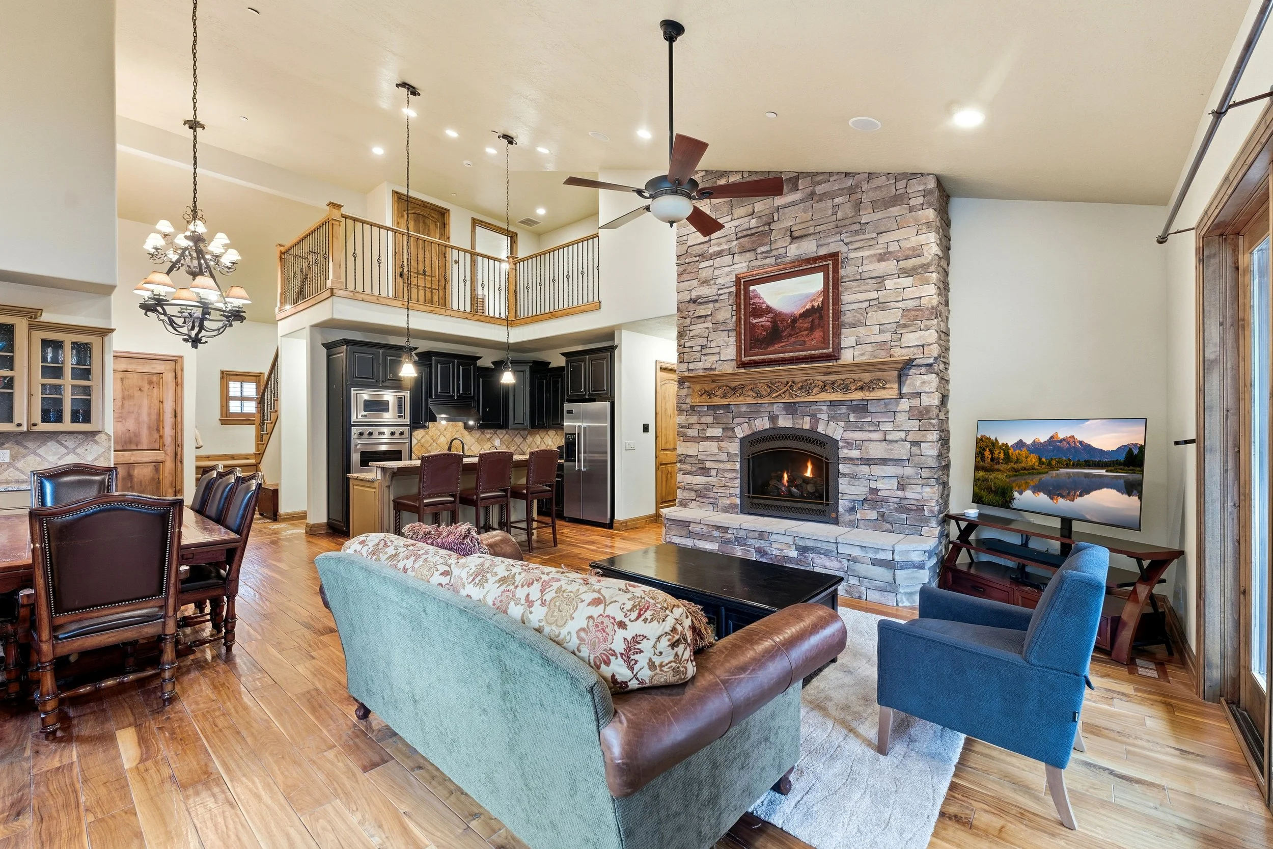 Spacious living room with a stone fireplace, ceiling fan, large window, flat-screen TV, dining area, and kitchen with black cabinets.