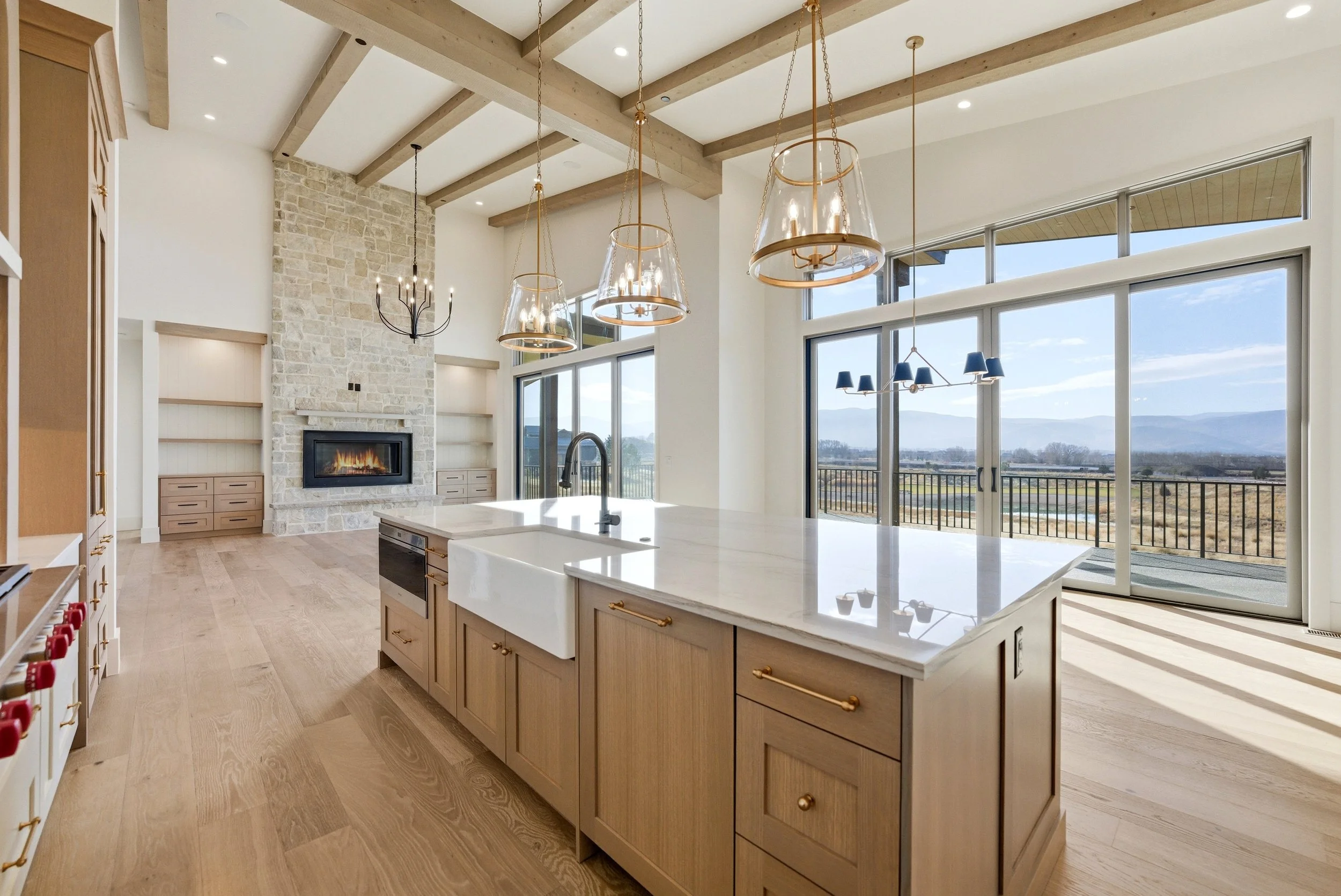 Modern kitchen with large island, wooden cabinets, and a view of mountains through floor-to-ceiling windows. Features hanging pendant lights and a stone fireplace.