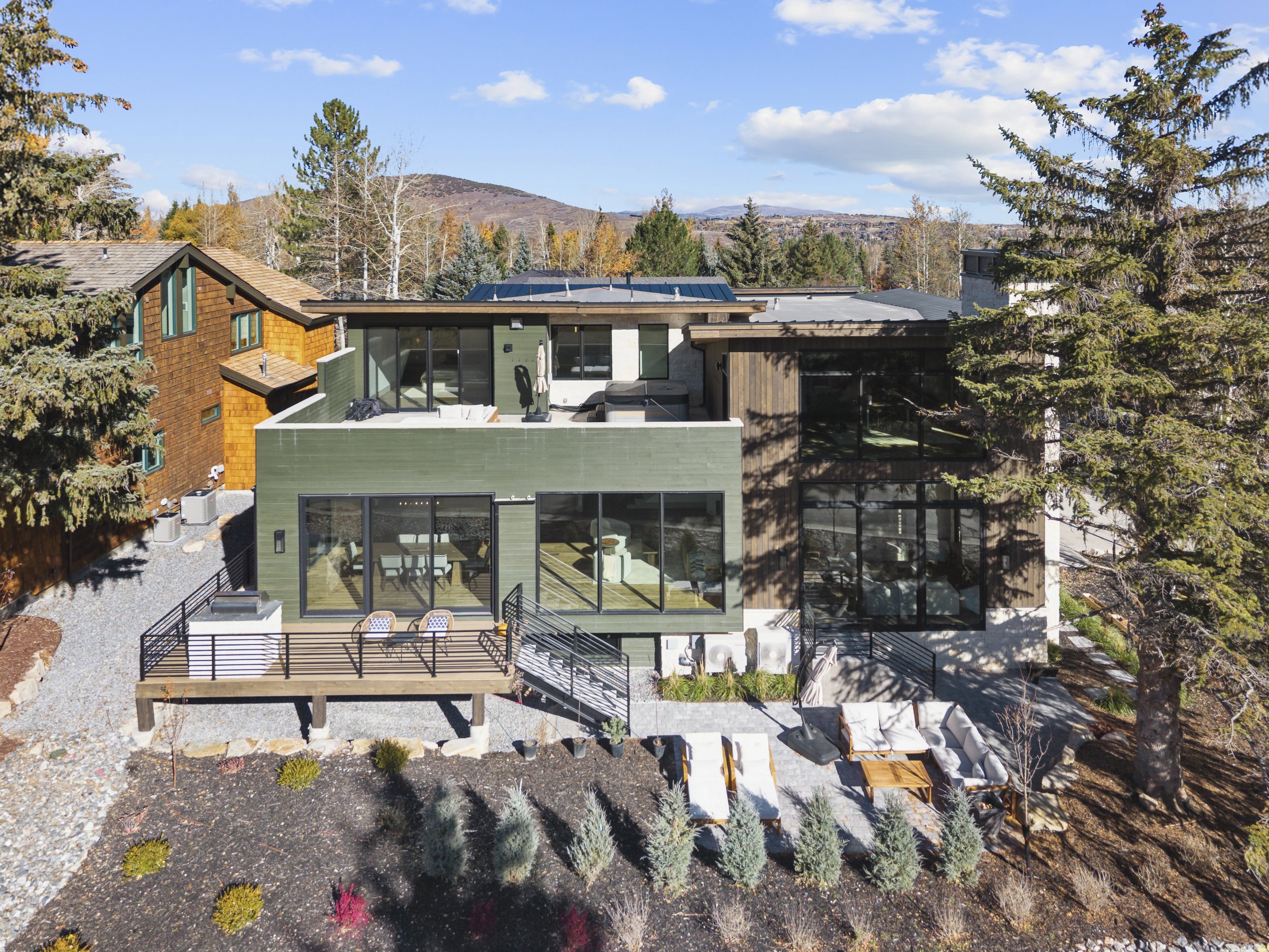 A modern multi-story house with large windows, surrounded by trees and a landscaped yard, with mountains and a blue sky in the background.