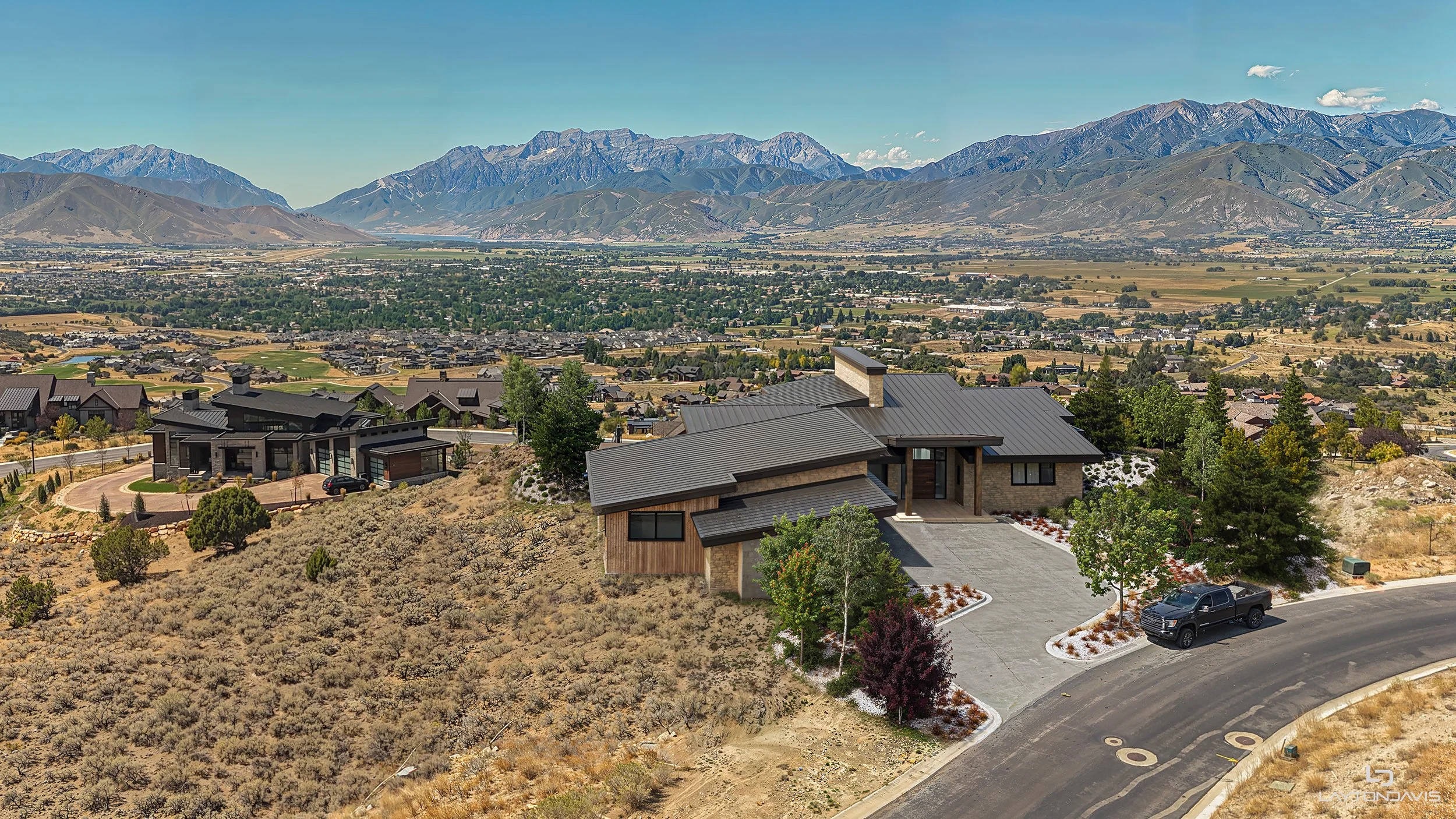 A modern house with a gray roof and a driveway, surrounded by trees and landscaping, overlooking a suburban area with mountains in the background.