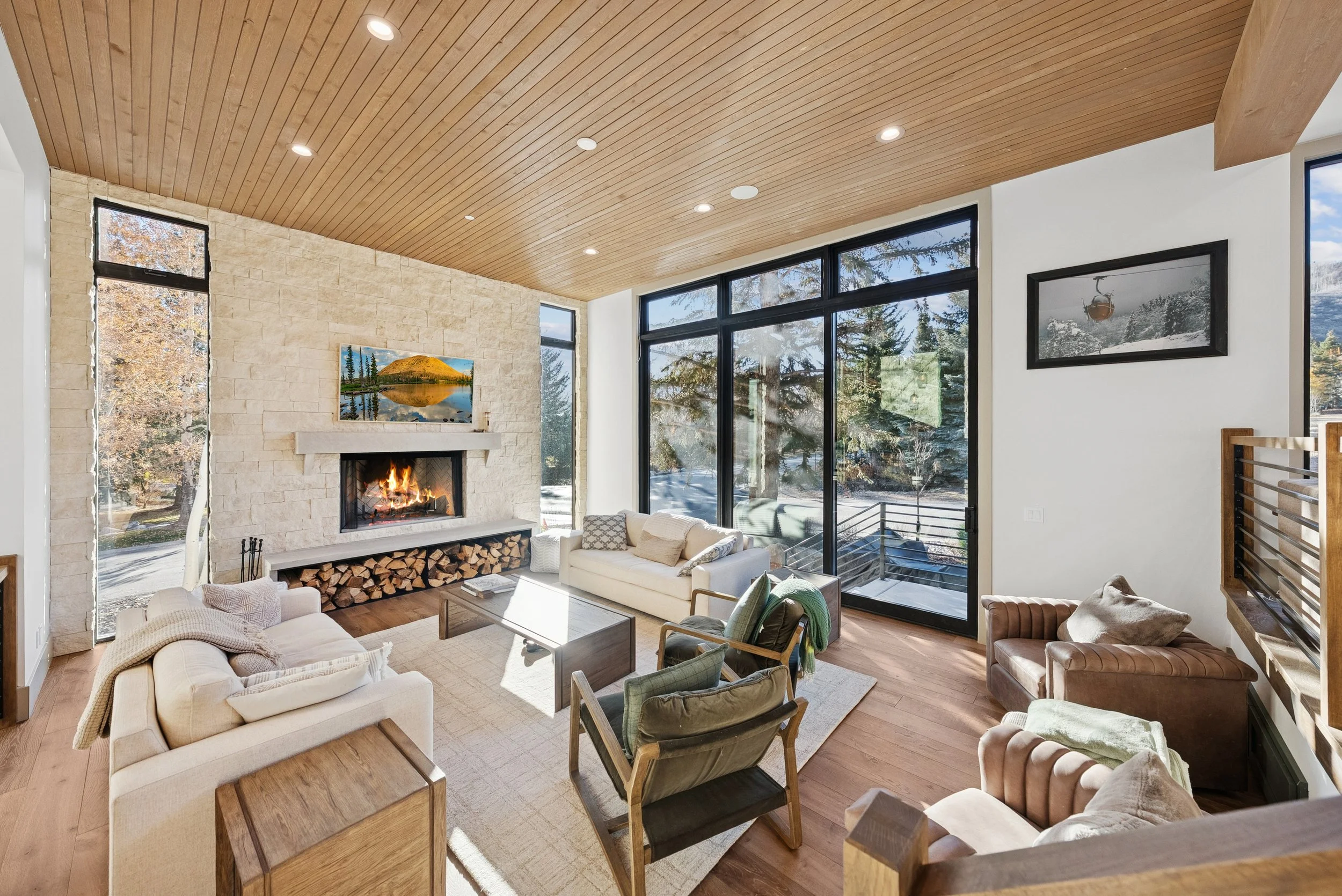 Modern living room with a wooden plank ceiling, large windows showing trees outside, a stone fireplace with a fire burning, and various sofas and chairs arranged around a central coffee table.
