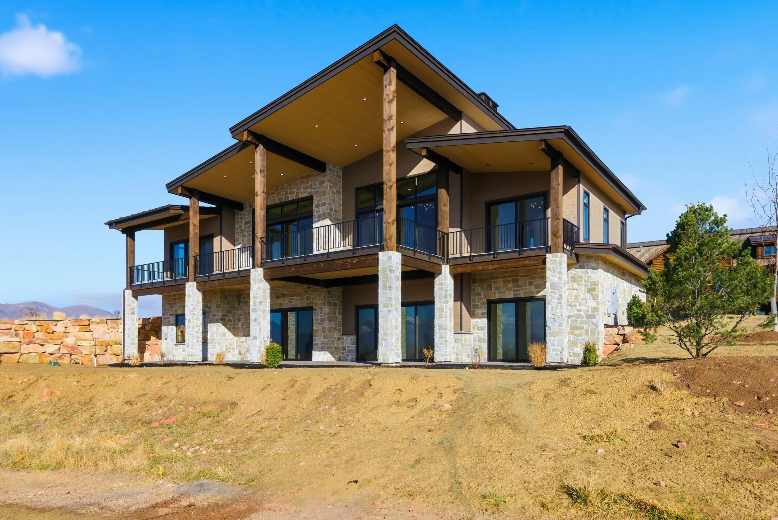 A modern house with a stone and wood exterior, large glass windows and a spacious balcony, situated on a grassy plot under a blue sky with some clouds.