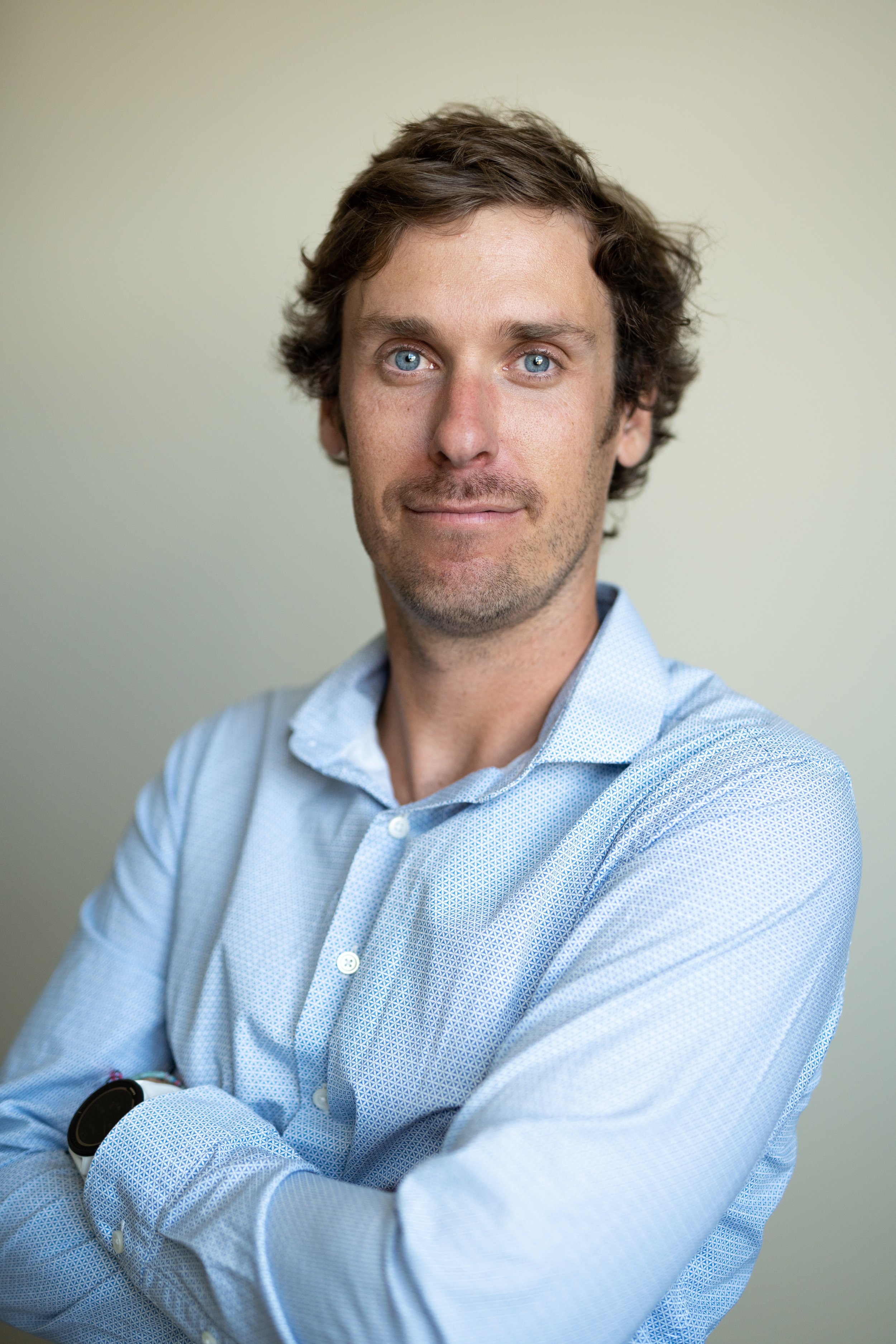 Headshot of a man with blue eyes and wavy brown hair, wearing a light blue patterned button-down shirt, standing against a plain, light-colored background.